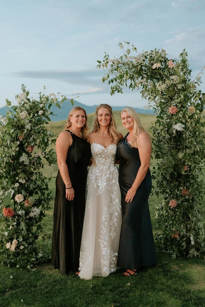 Three women standing in front of a floral wedding arch outdoors, with green hills and mountains in the background, during daytime.