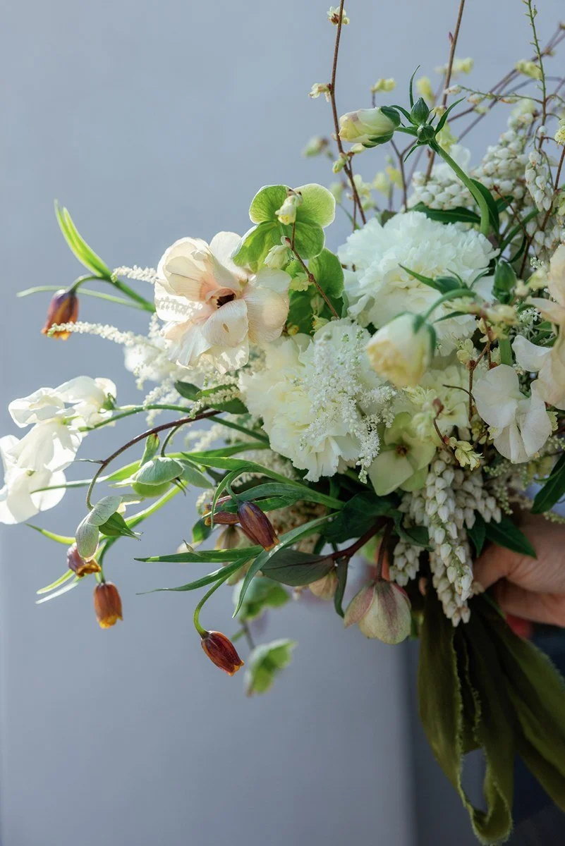 Assorted colorful flower bouquet with roses, daisies, and greenery on a dark table against a plain wall.