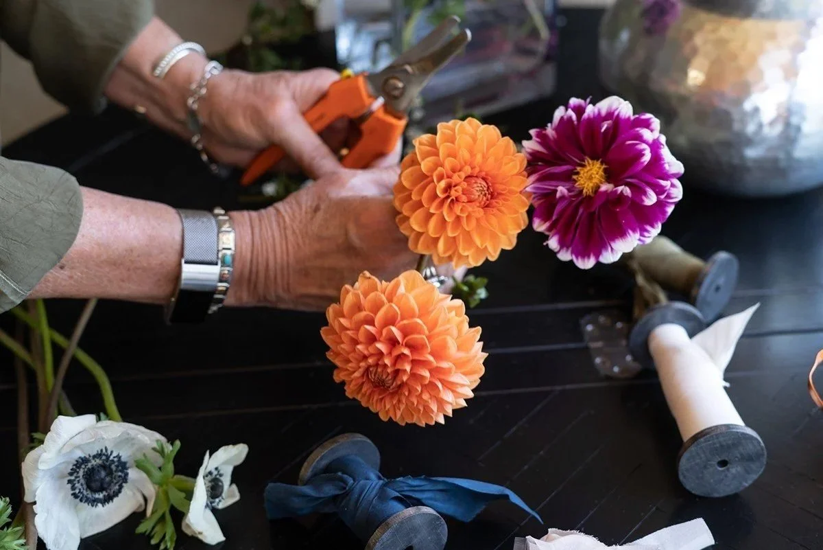 Person arranging bright orange and purple flowers at a floral arrangement table, using pruning shears. The table has rolls of wire, ribbon, and flowers.