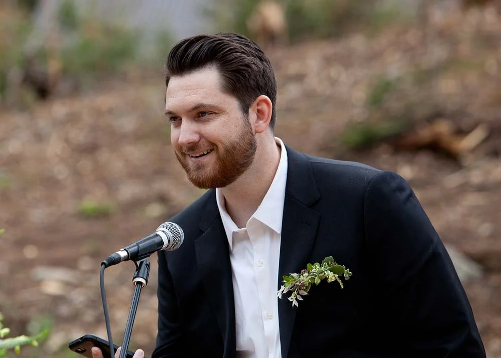 A man in a black suit and white shirt speaking into a microphone outdoors, with a boutonniere on his lapel.