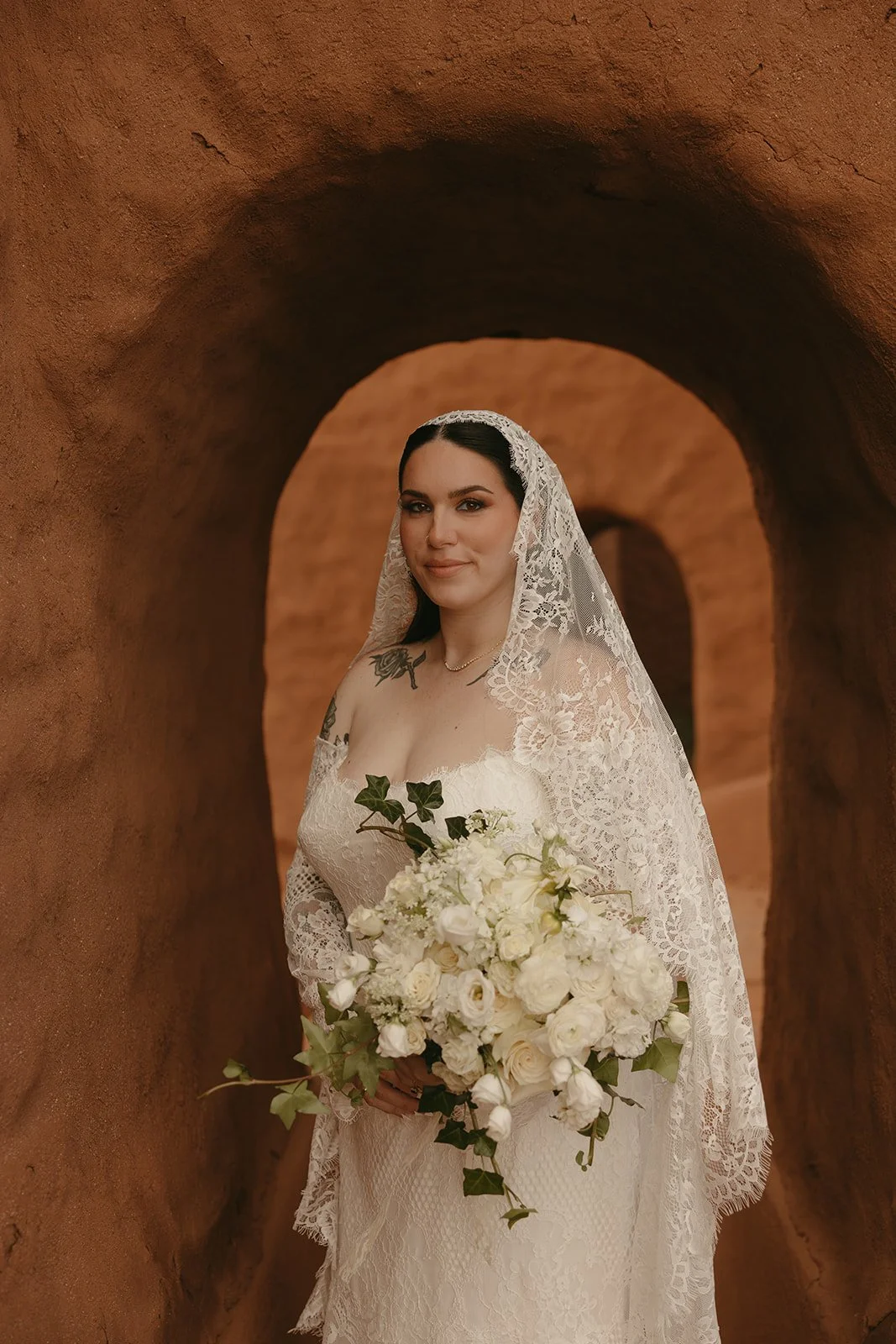 A bride in a lace wedding dress holding a bouquet of white flowers, standing in a brown stone archway.