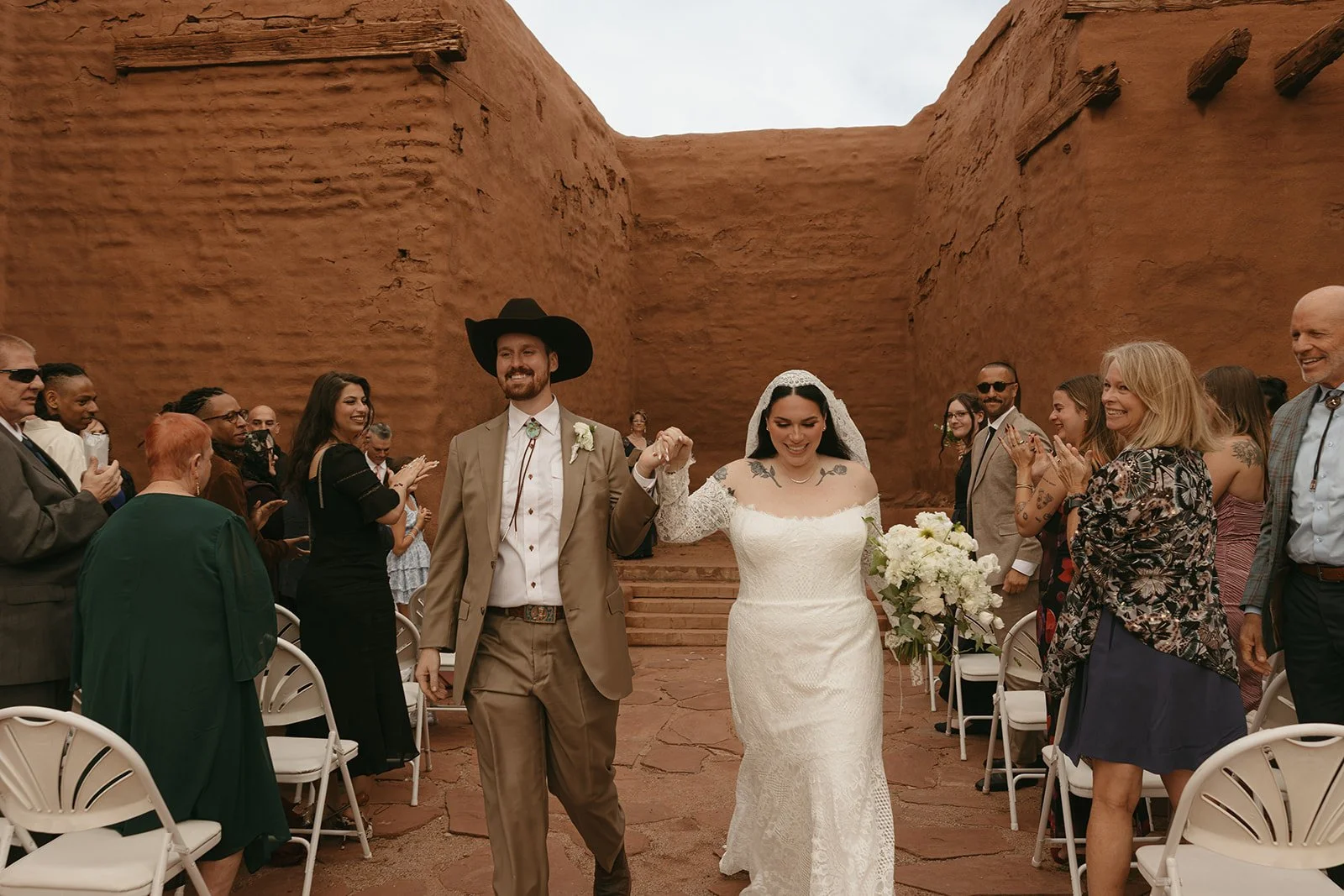 A wedding couple holding hands and walking down the aisle in an outdoor desert setting with red rock formations, surrounded by seated guests clapping and smiling.