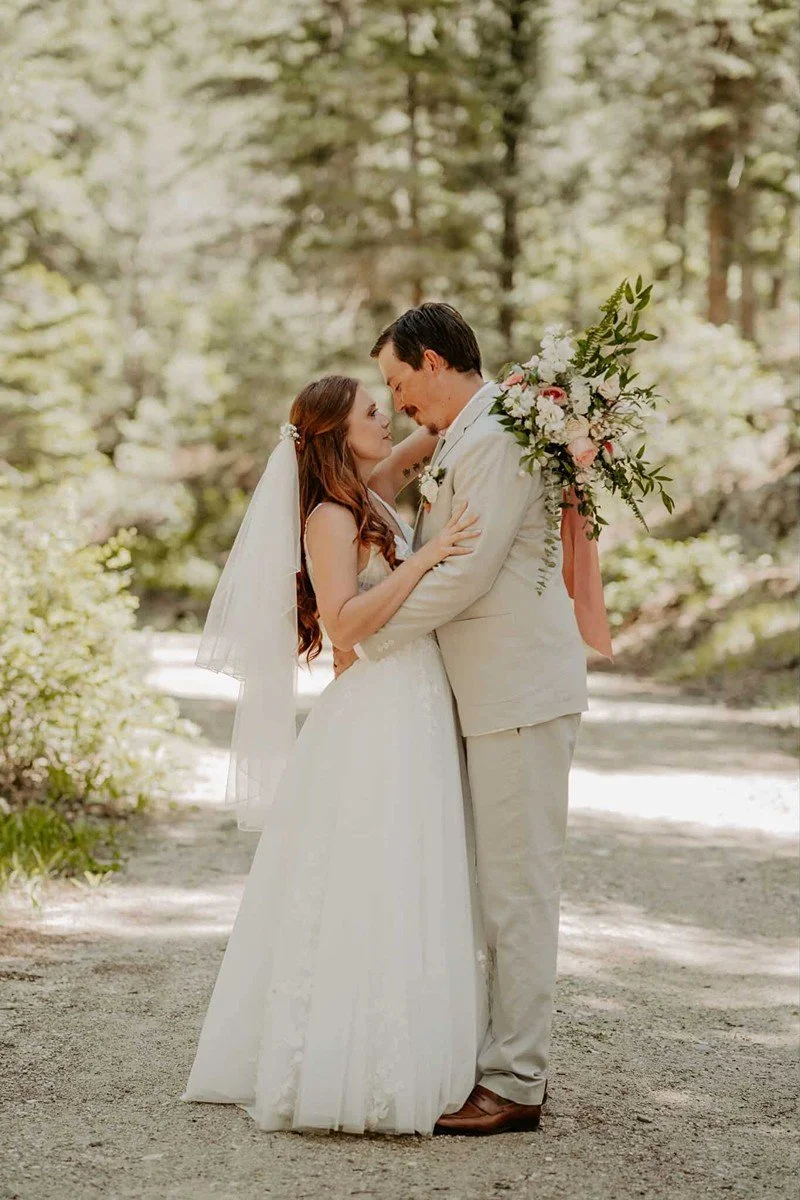A bride and groom standing close together outdoors in a wooded area, gazing into each other's eyes. The bride is wearing a white wedding dress and veil, and the groom is dressed in a light-colored suit, holding a bouquet of flowers over his shoulder.