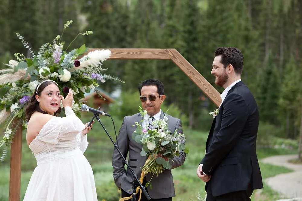 A bride is crying while holding a microphone during her outdoor wedding, with a groom smiling in front of her and an officiant holding a bouquet, surrounded by a wooden arch and greenery.