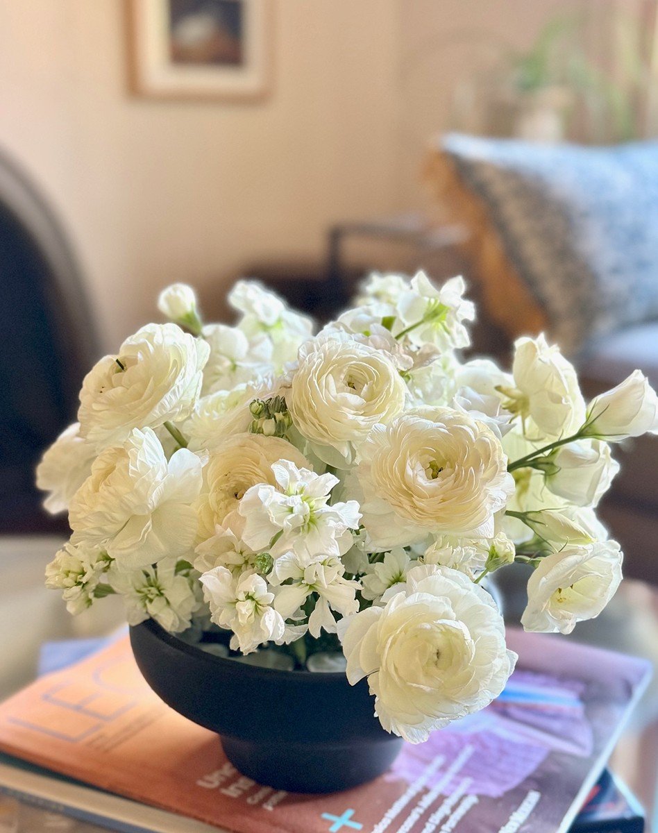 A black bowl filled with white flowers, placed on top of magazines and papers on a table, with a living room background.