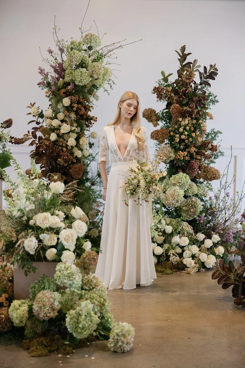 A woman in a white dress surrounded by large flower arrangements and floral displays, standing indoors against a plain white wall.