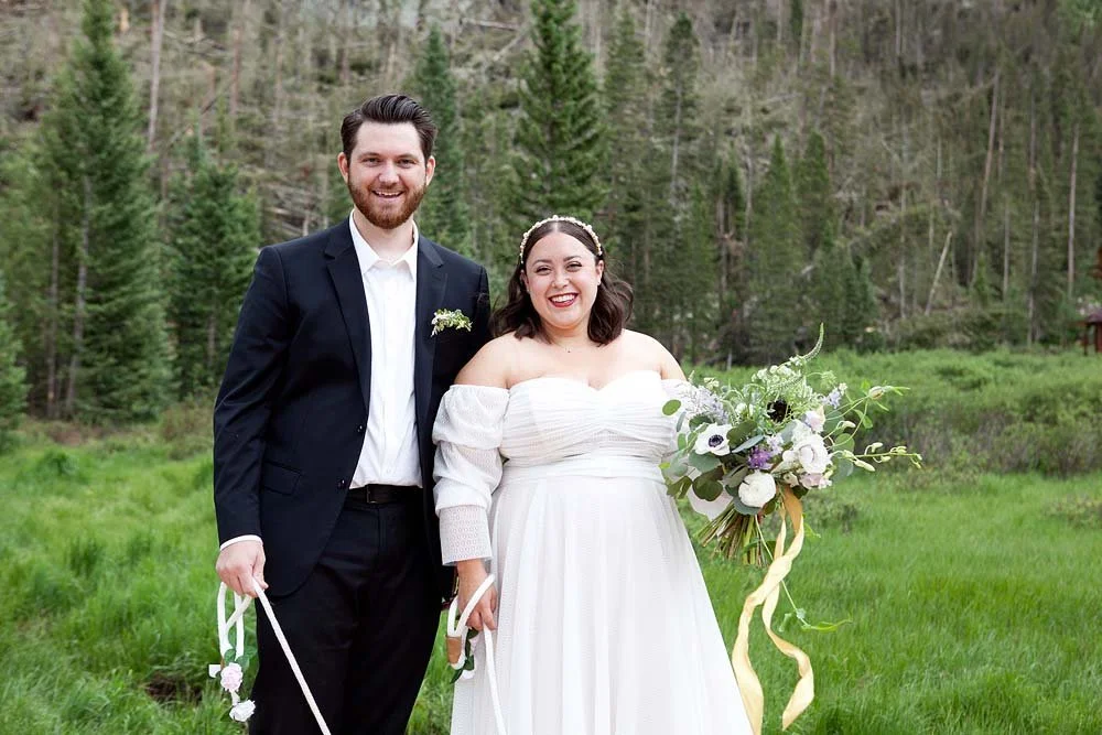 A smiling bride and groom stand together outdoors in a green field with trees in the background, during a wedding. The bride wears a white off-the-shoulder wedding dress and holds a bouquet of white and purple flowers. The groom wears a dark suit wit