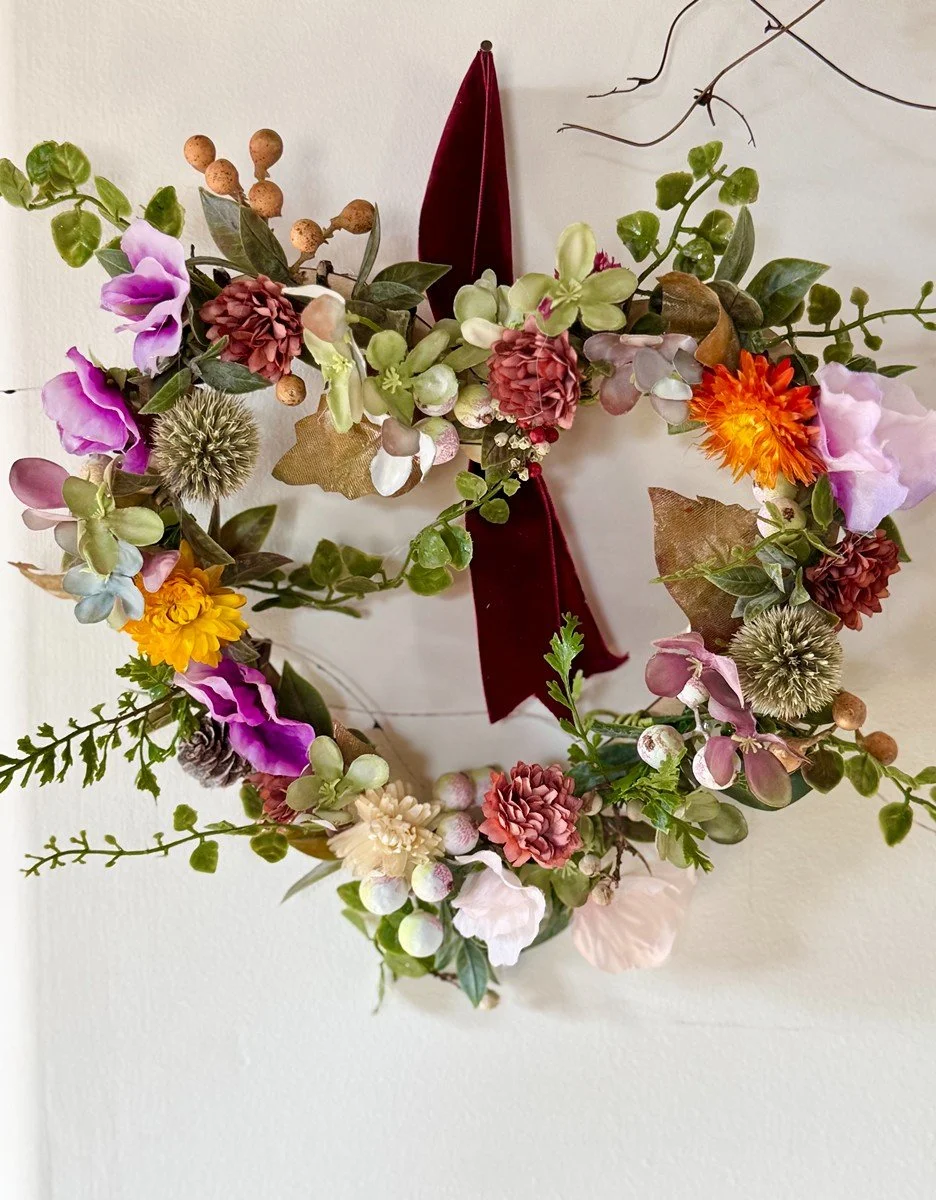 Floral wreath with pink, purple, yellow, white, and orange flowers, greenery, and a red ribbon, hung on a white wall.