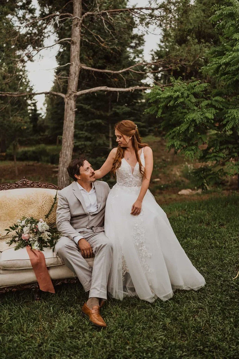 A bride and groom outdoors in a wooded area, with the bride standing and the groom sitting on a vintage sofa. The groom is dressed in a light gray suit and brown shoes, while the bride wears a white wedding dress. There is a bouquet of flowers on the sofa, and the scene is set in a natural, green environment.