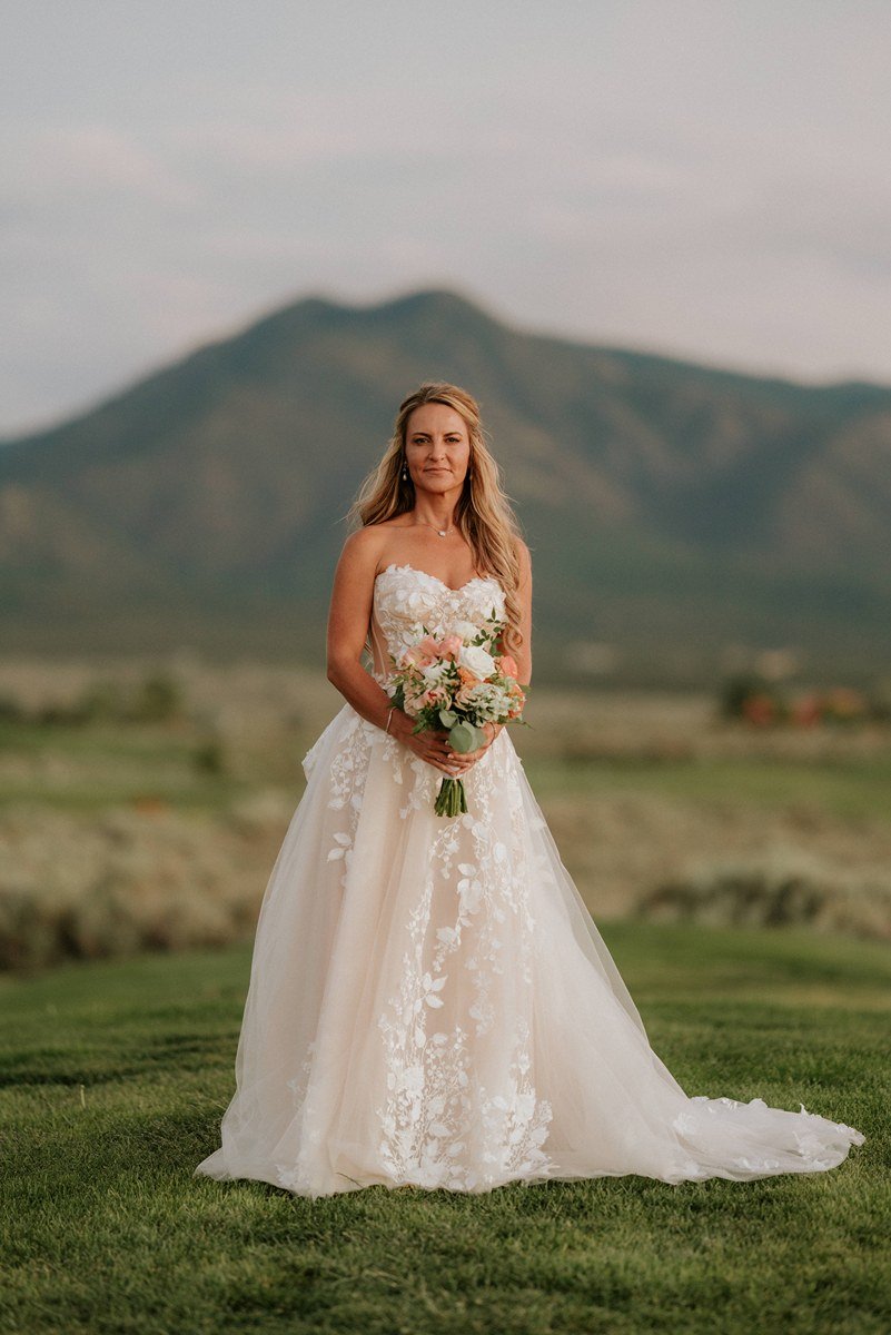 A woman in a strapless wedding dress holding a bouquet of flowers, standing outdoors with mountains in the background.
