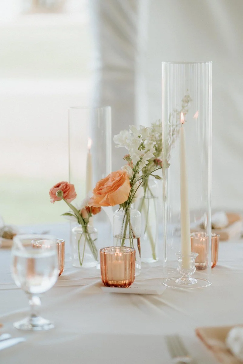 A table centerpiece with white and peach flowers in clear glass vases, surrounded by pink and clear candle holders, on a white tablecloth with candles and dishware.
