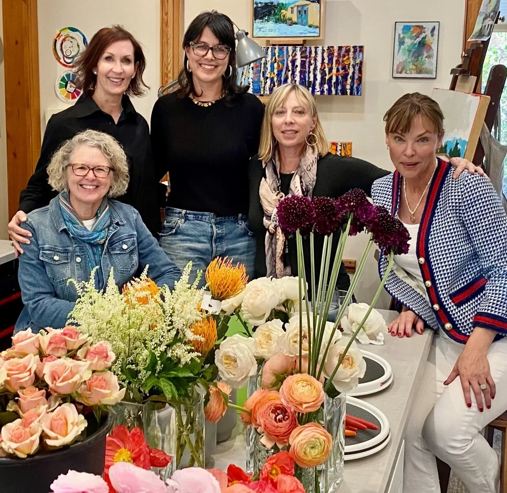 Five women gathered around a table filled with colorful flowers, in an art studio with paintings on the wall.