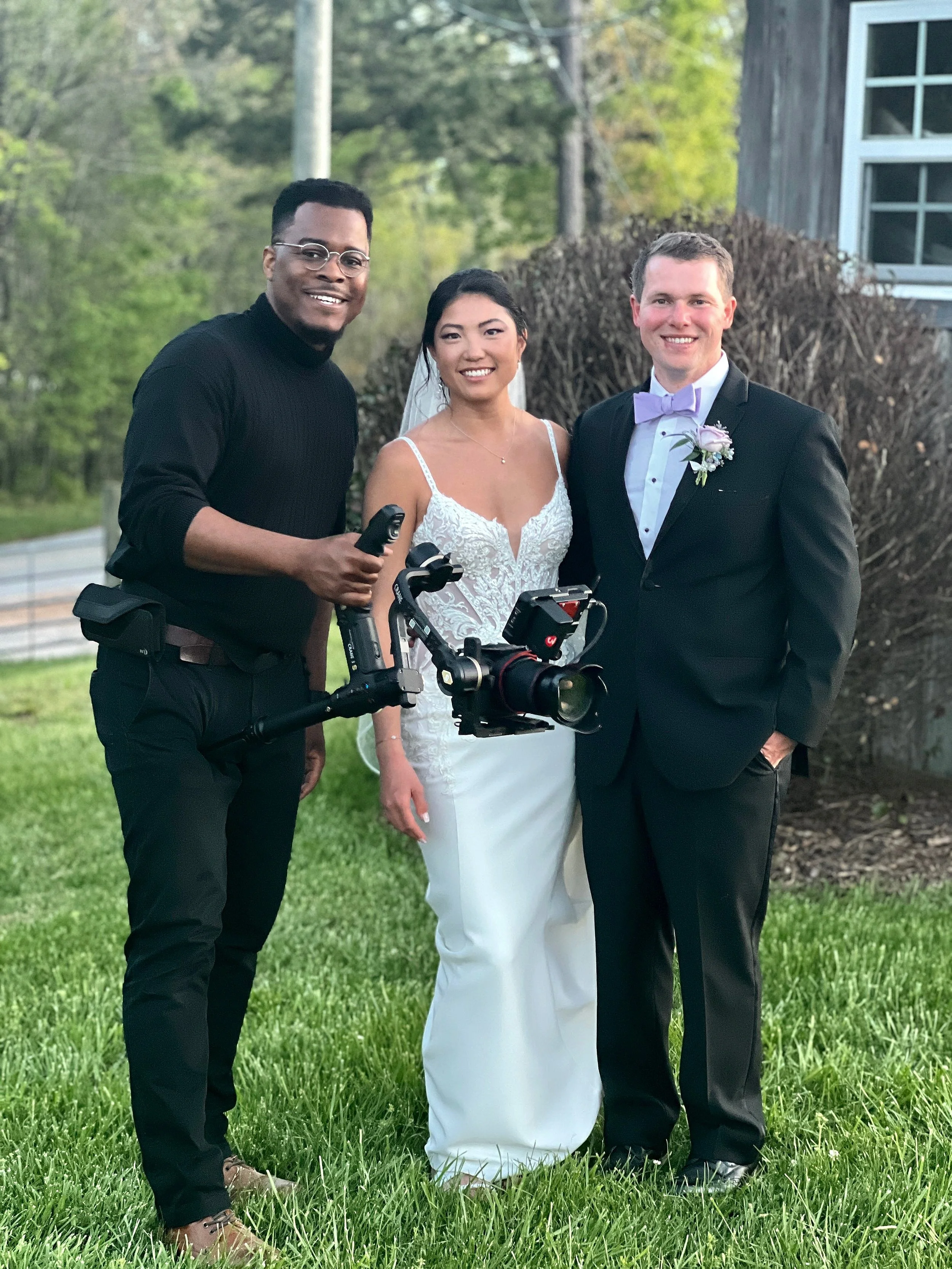 A smiling bride and groom pose with a videographer holding a camera on a stabilizer, standing outdoors on grass in front of some bushes.