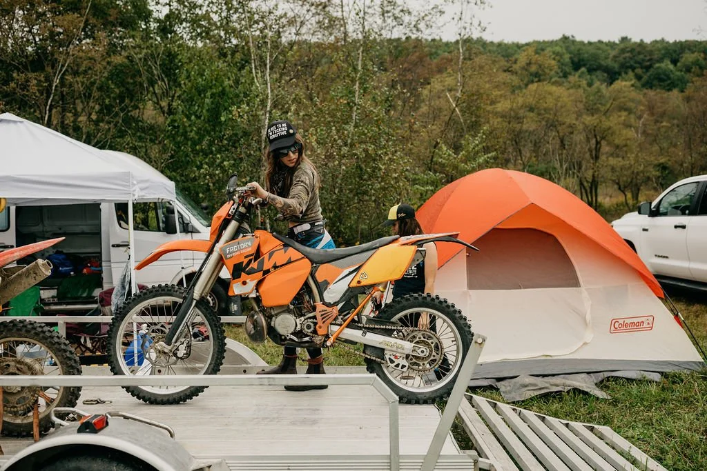 A woman is working on a KTM dirt bike on a trailer, with a tent, vehicles, and trees in the background during a camping trip.