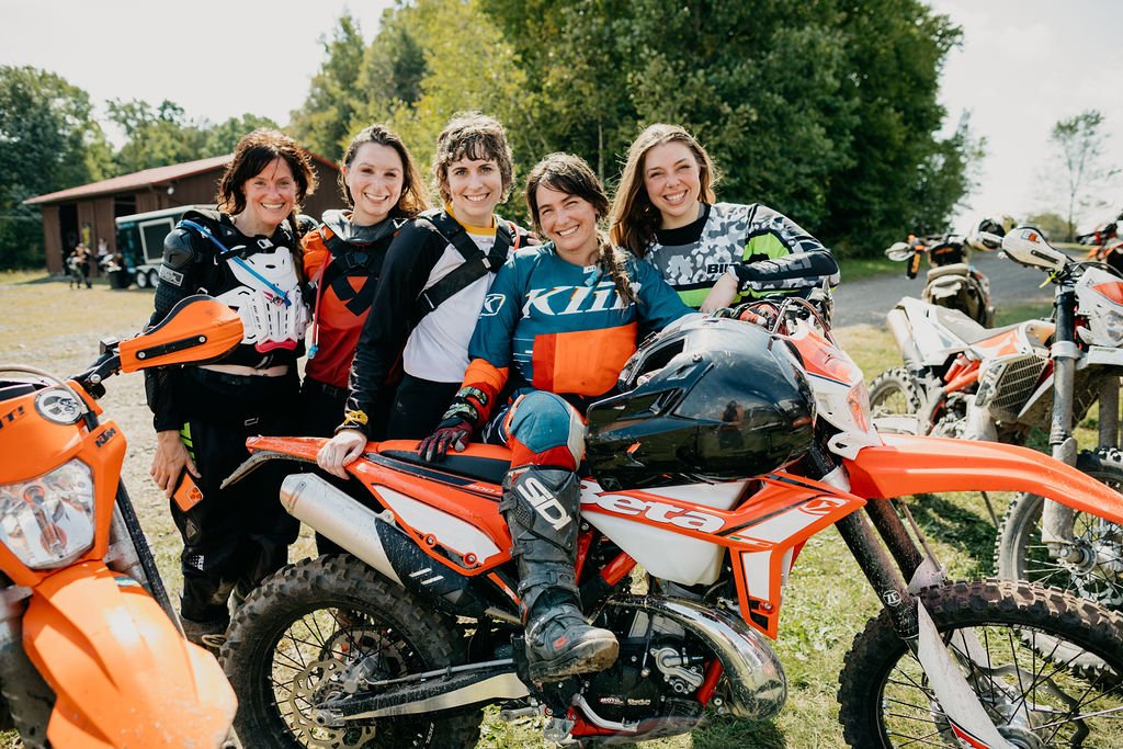 Group of five women in motocross gear smiling, with dirt bikes, outdoors on a grassy field with trees in the background.