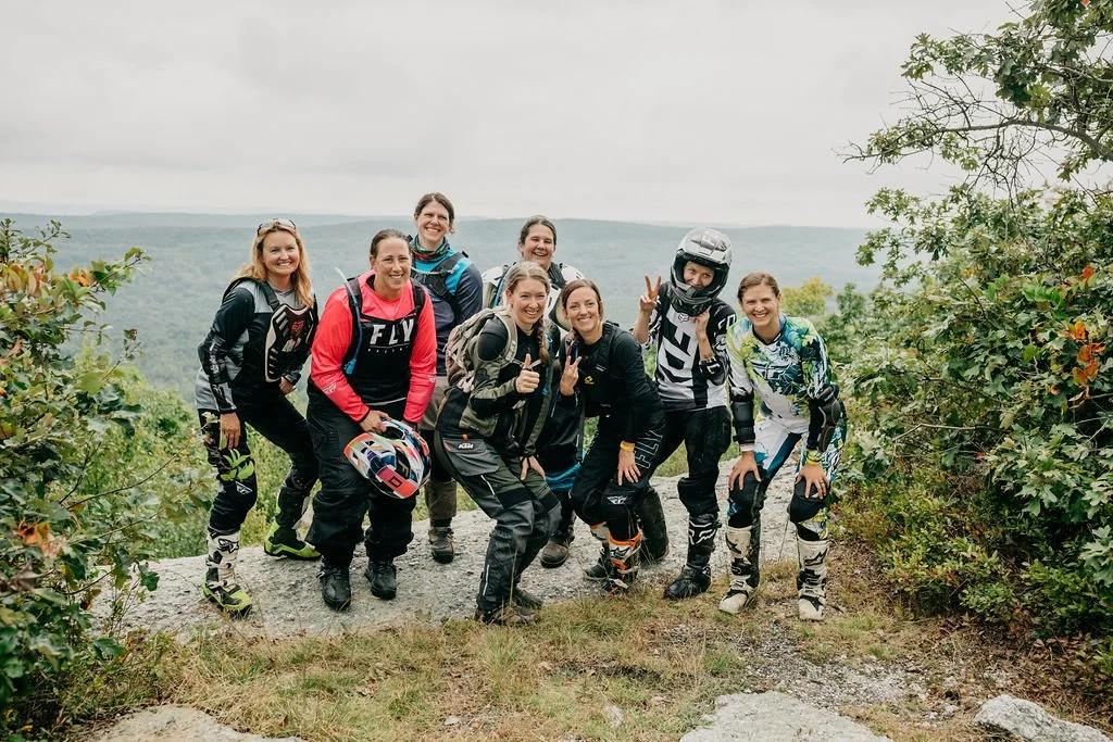 A group of eight women in motocross gear, posing outdoors on a trail with greenery and hills in the background.