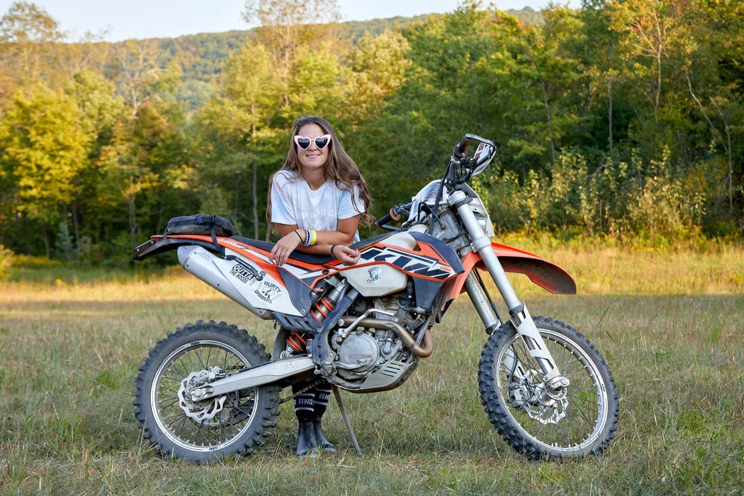 A young woman with long brown hair wearing sunglasses and a white t-shirt, leaning on an orange KTM dirt bike, standing in a grassy field with trees and hills in the background.