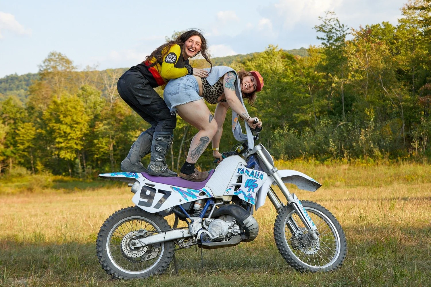 Two women riding a dirt bike outdoors in a field with trees in the background. The woman in front is leaning forward, wearing a leopard print top, shorts, and a red cap, while the woman behind is smiling and wearing a yellow and black jacket with bla