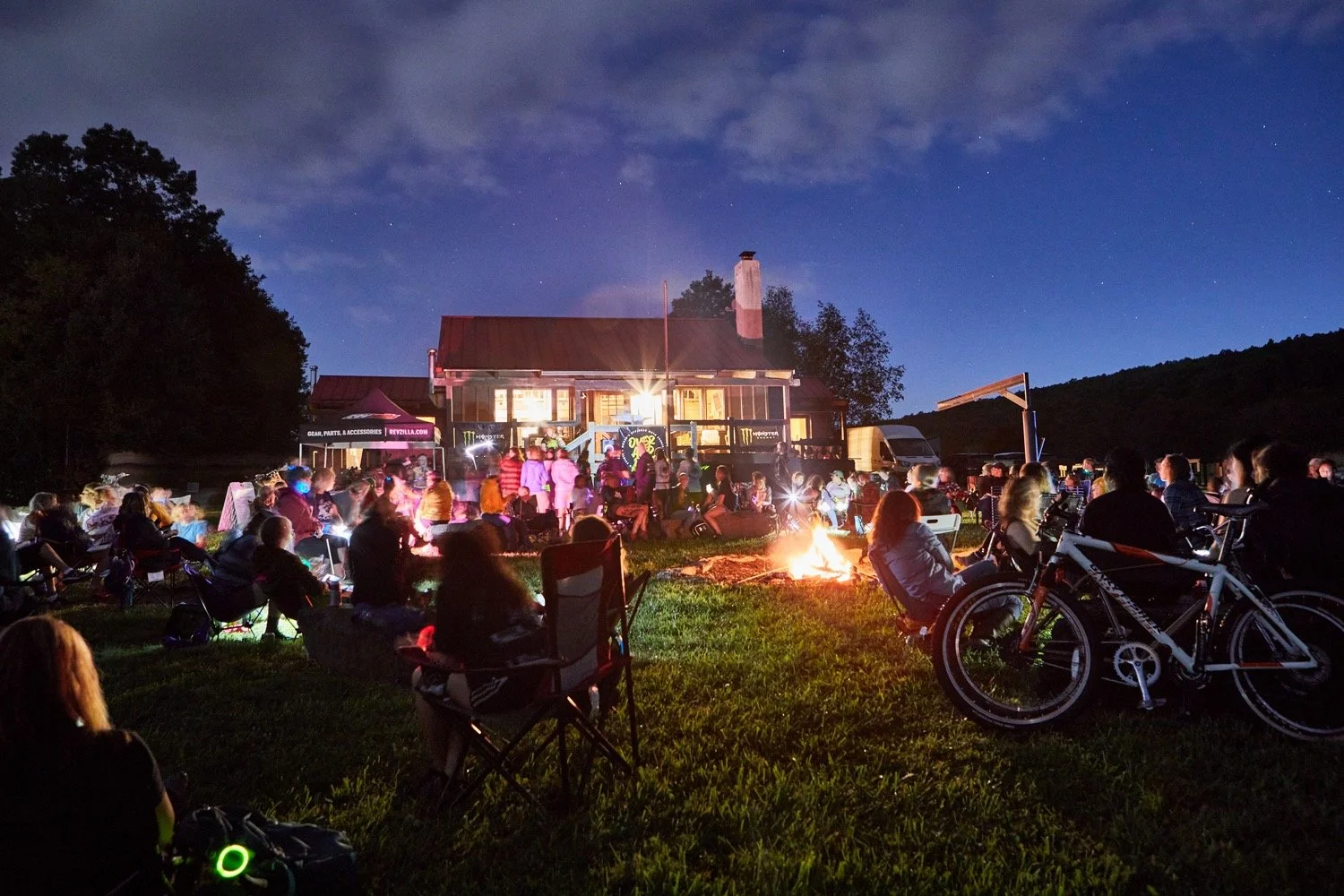People gathered outdoors around a campfire at night, some sitting in chairs, others standing, in front of a rustic house with lit windows, under a starry sky, with bicycles and camping gear nearby.