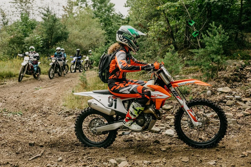 A woman wearing motocross gear and a helmet riding an orange dirt bike on a rocky trail with a group of people on bikes following behind in a green, forested area.
