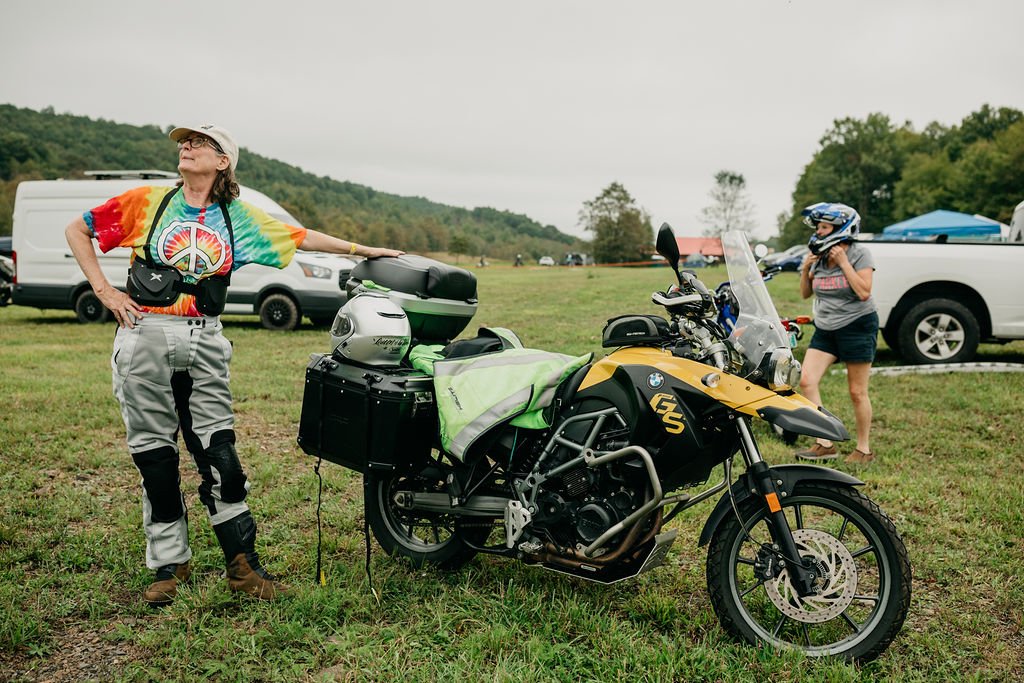 A woman in colorful tie-dye shirt and motorcycle gear stands with one hand on a black and yellow BMW motorcycle. A young girl wearing a helmet stands beside a white pickup truck in a grassy field with other vehicles and trees in the background.