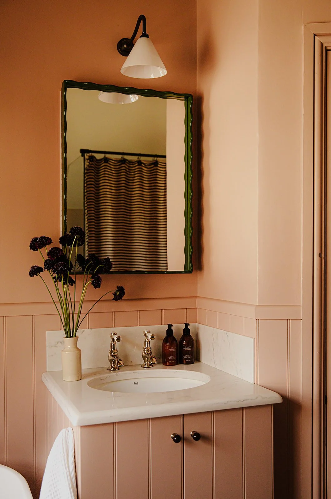 Bathroom with pink wall paneling, marble countertop sink, mirror with green frame, striped shower curtain visible in reflection, white vase with dark purple flowers, two brown soap dispensers.