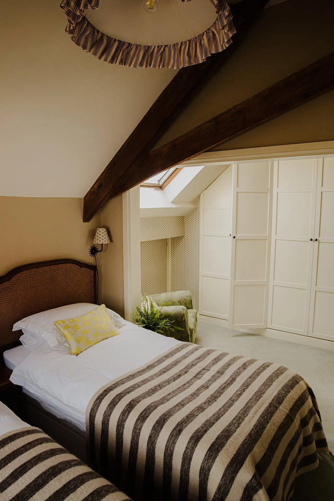 A cozy bedroom with a bed, striped blanket, headboard, and a yellow pillow. There is a wall-mounted lamp next to the bed, a large white built-in closet, a plush armchair near a skylight window, and a potted fern plant. The room has sloped ceiling with exposed wooden beams.