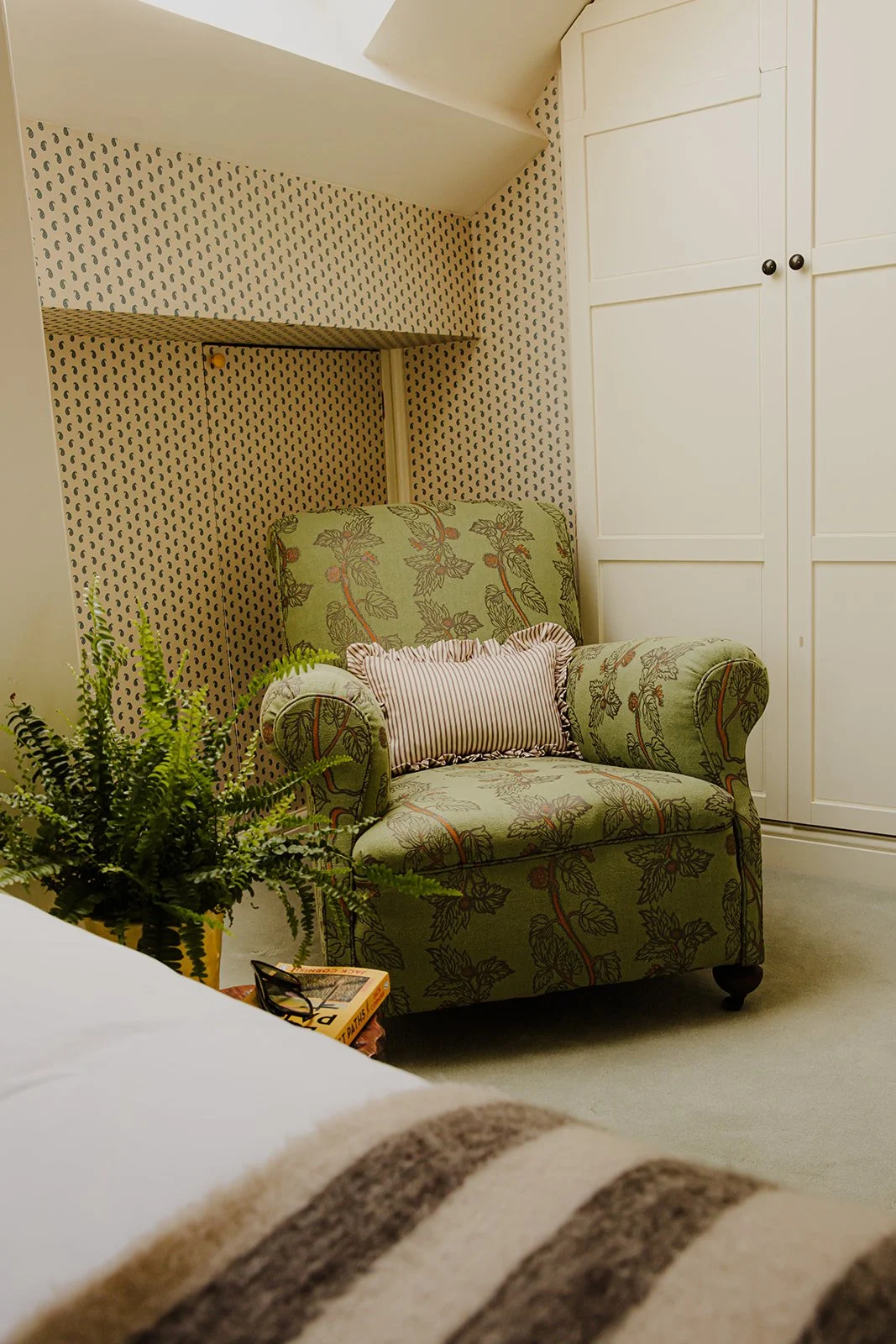 A cozy corner with a floral upholstered armchair, a cushion, a potted fern, a pair of glasses on a book, and a white closet with black knobs in the background.