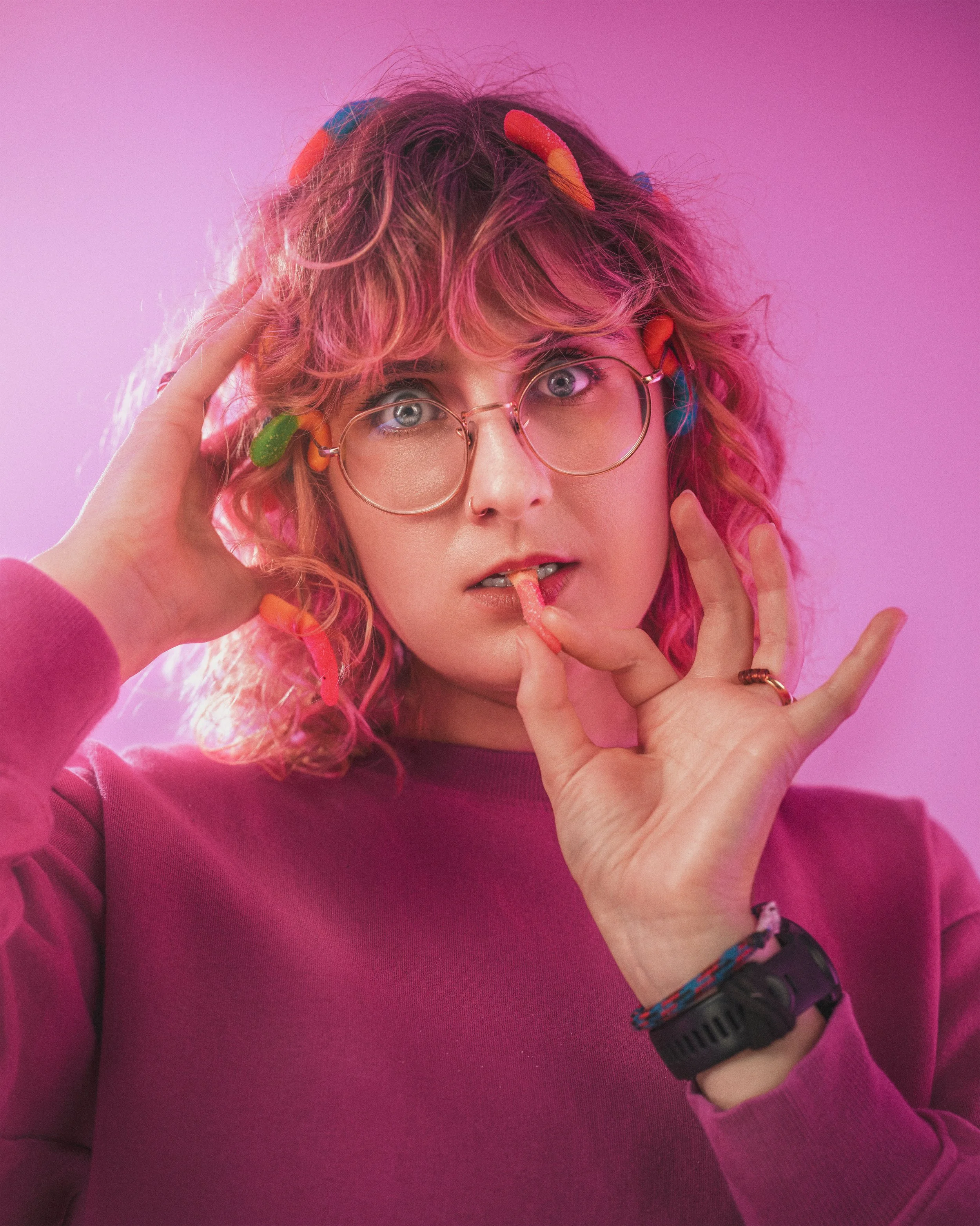 Young woman with curly pink hair wearing glasses and a pink sweater, surrounded by colorful gummy worms, holding one gummy worm to her lips, with a pink background.