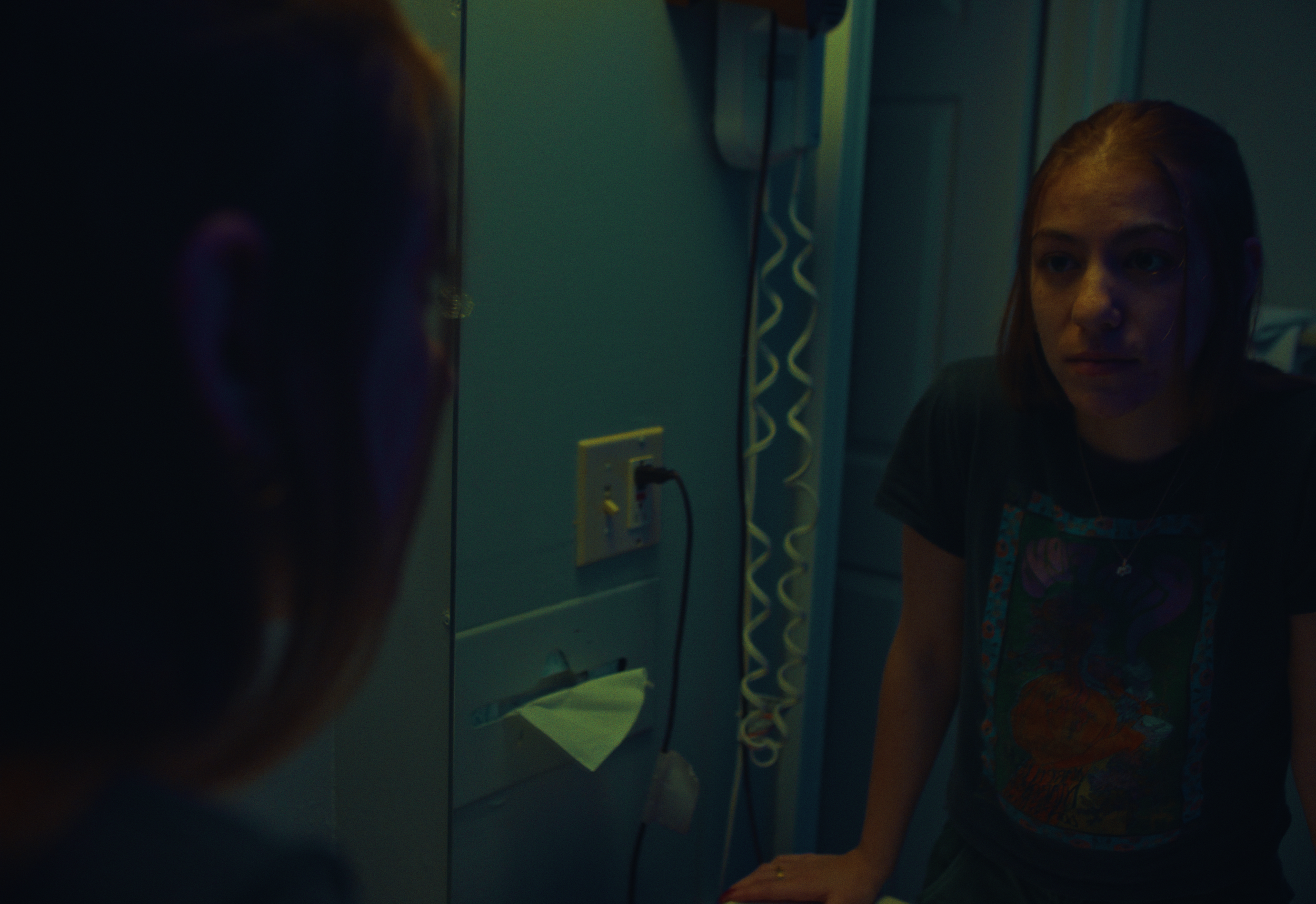 A young woman with shoulder-length hair looking at her reflection in a mirror, with dim lighting in a bathroom. The bathroom has a wall socket with a plug and a yellow paper towel holder.