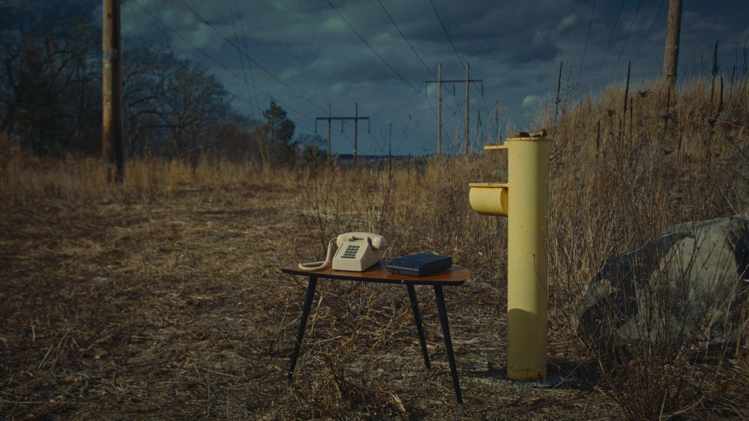 An outdoor scene featuring a yellow telephone booth, a small wooden table with a vintage rotary phone and a closed portable electronic device, dry grass, a large rock, and power lines, under a cloudy sky.