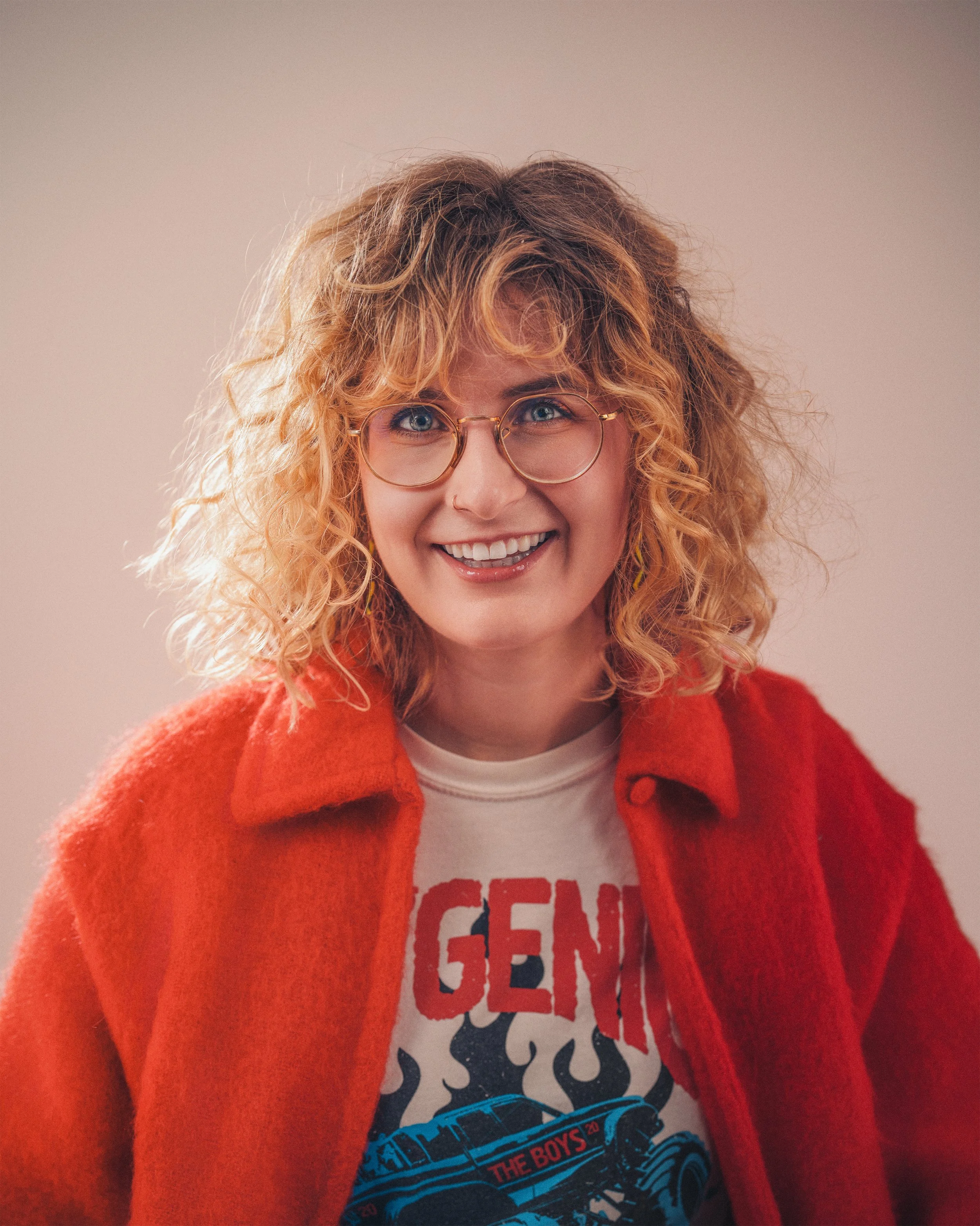 A woman with curly blonde hair and glasses smiles, wearing a red jacket over a graphic T-shirt.