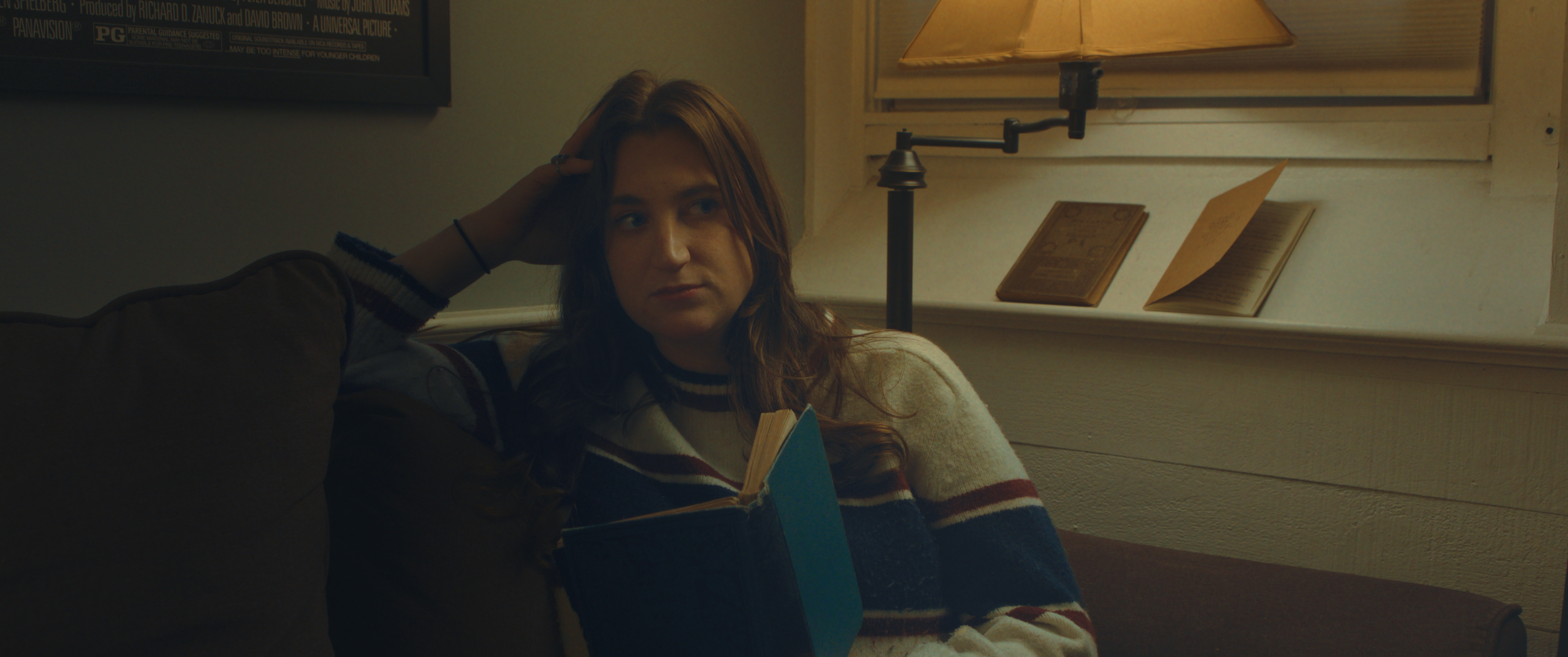 A young woman with long brown hair sitting on a sofa, holding an open book, looking thoughtfully to the side. The setting appears cozy with a lamp and window in the background.