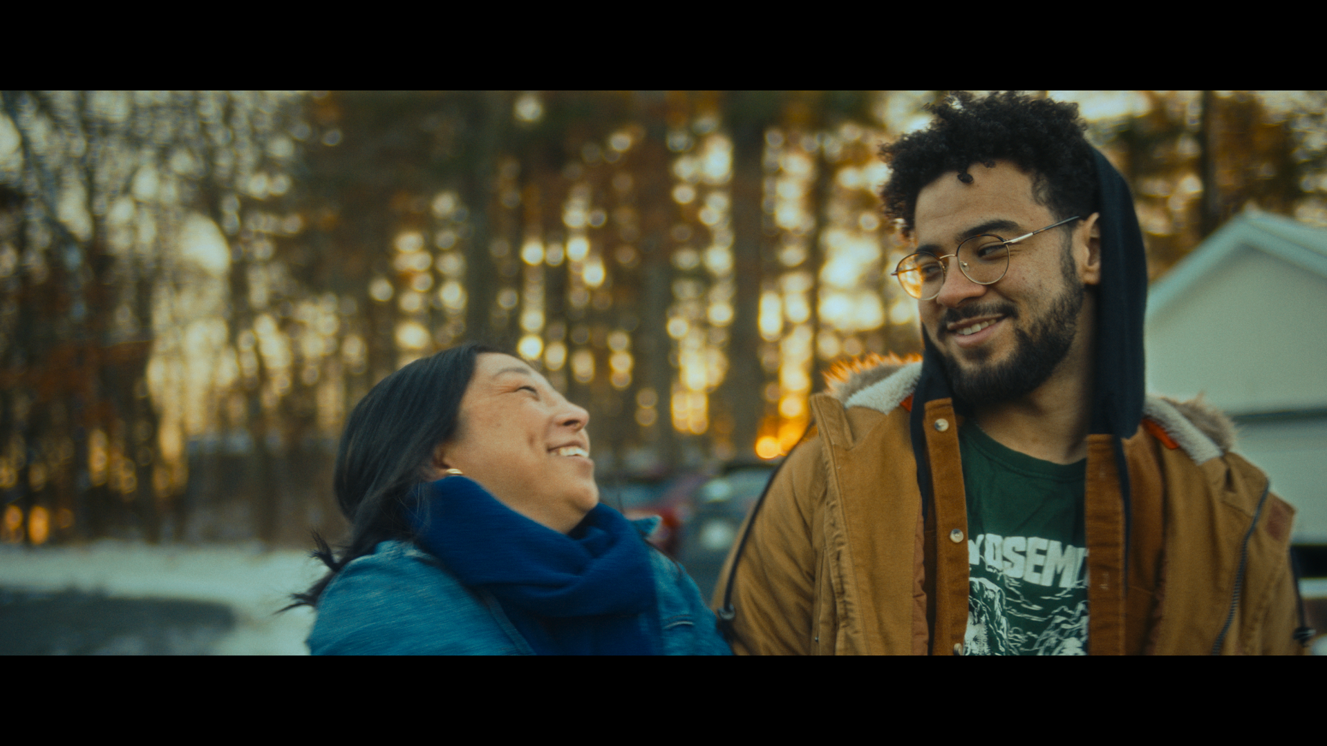 Two people smiling warmly at each other outdoors during sunset, with trees and parked cars in the background.
