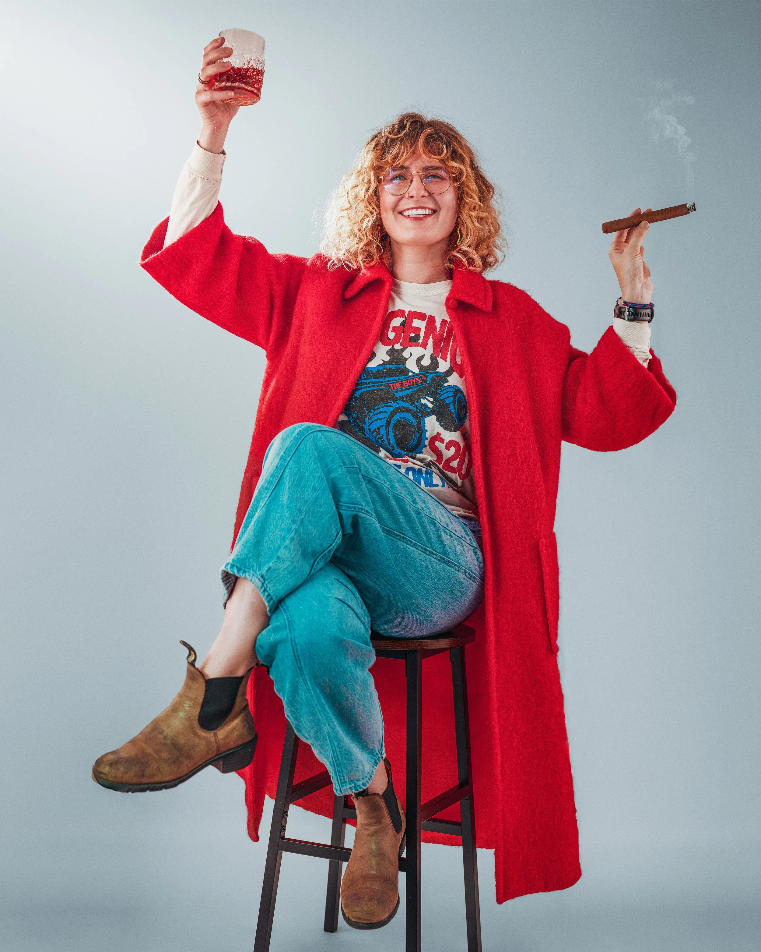 A woman with curly blonde hair, wearing glasses, a red coat, a graphic t-shirt, blue jeans, and brown ankle boots, sitting on a high stool, holding a drink in one hand and a cigar in the other, smiling.