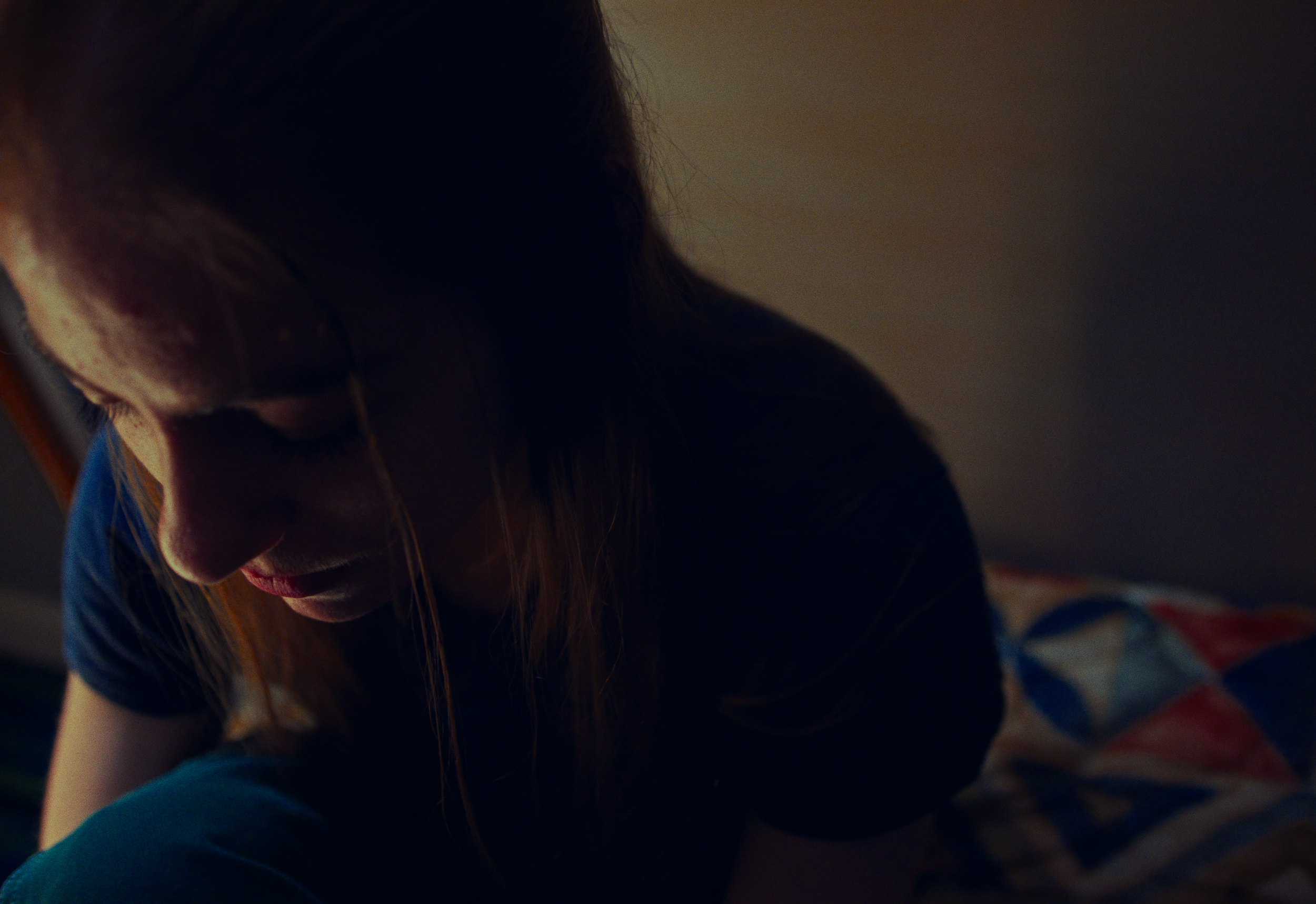 A young woman with red hair is sitting with her head bowed down in a dimly lit room, wearing a dark shirt and jeans.