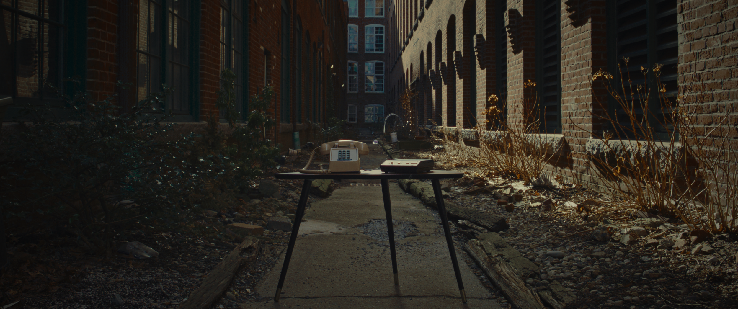 An abandoned outdoor alley with overgrown plants, dirt, and debris, featuring a table with an old rotary telephone and a vintage radio.