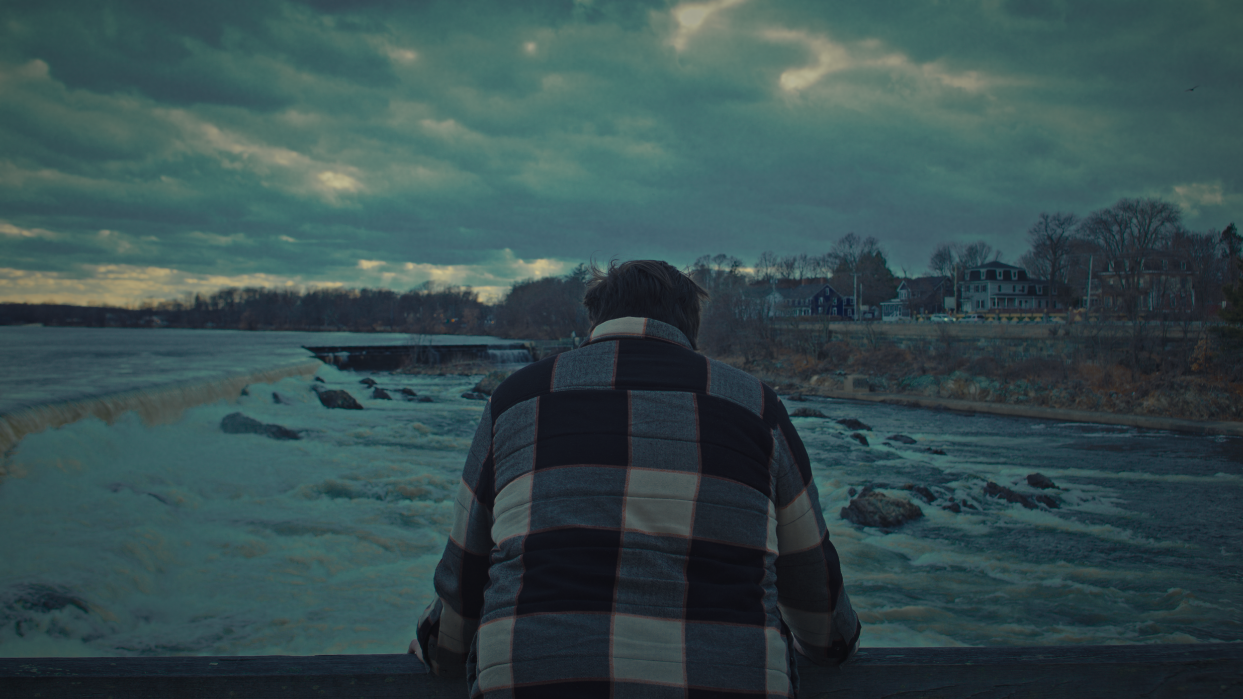 A person in a checkered jacket sits on a dock facing a river or waterfall with cloudy skies overhead.