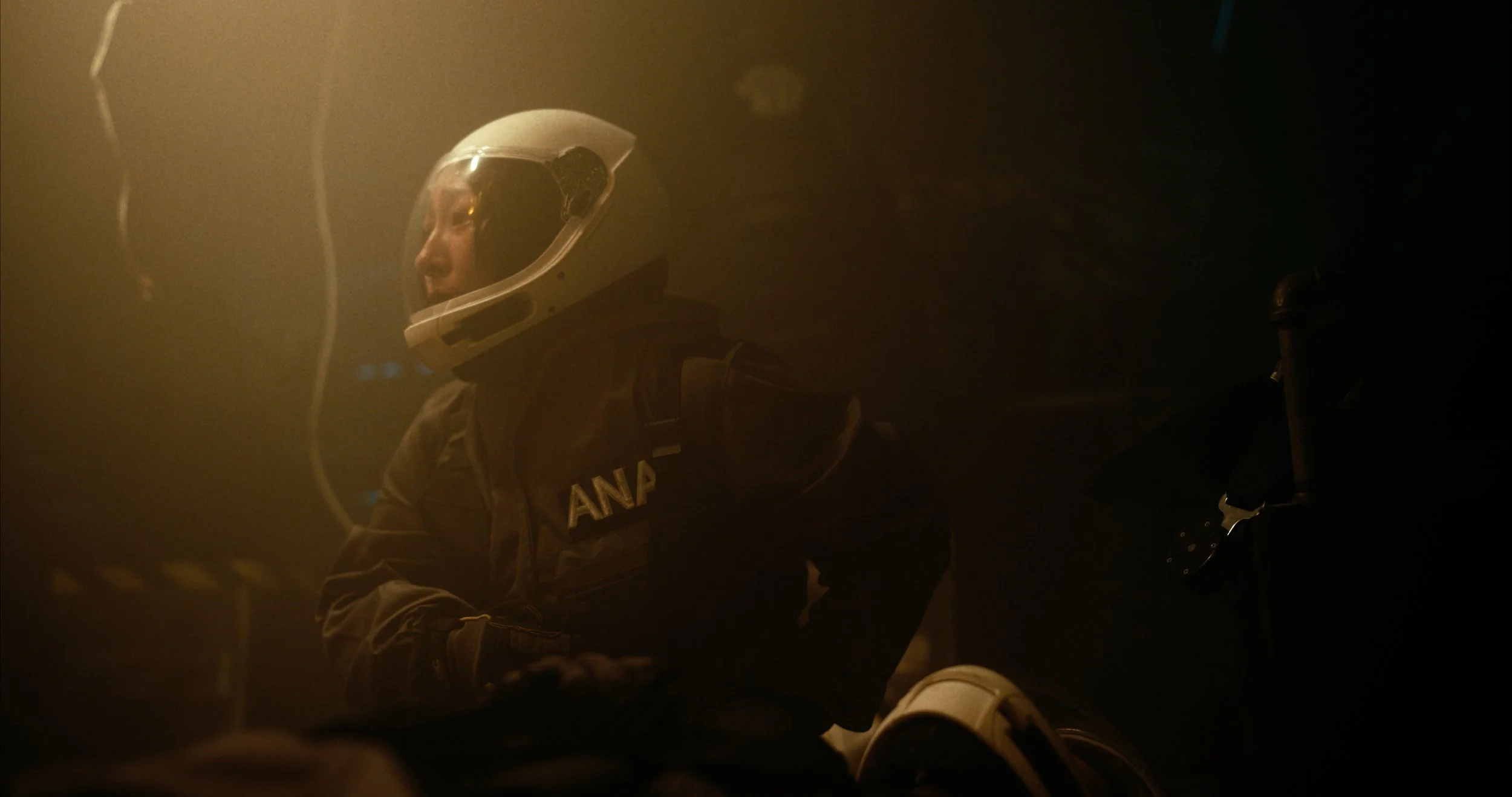 A person wearing an astronaut helmet and a jacket with 'ANA' on it, in a smoky or hazy environment, with a dark background.
