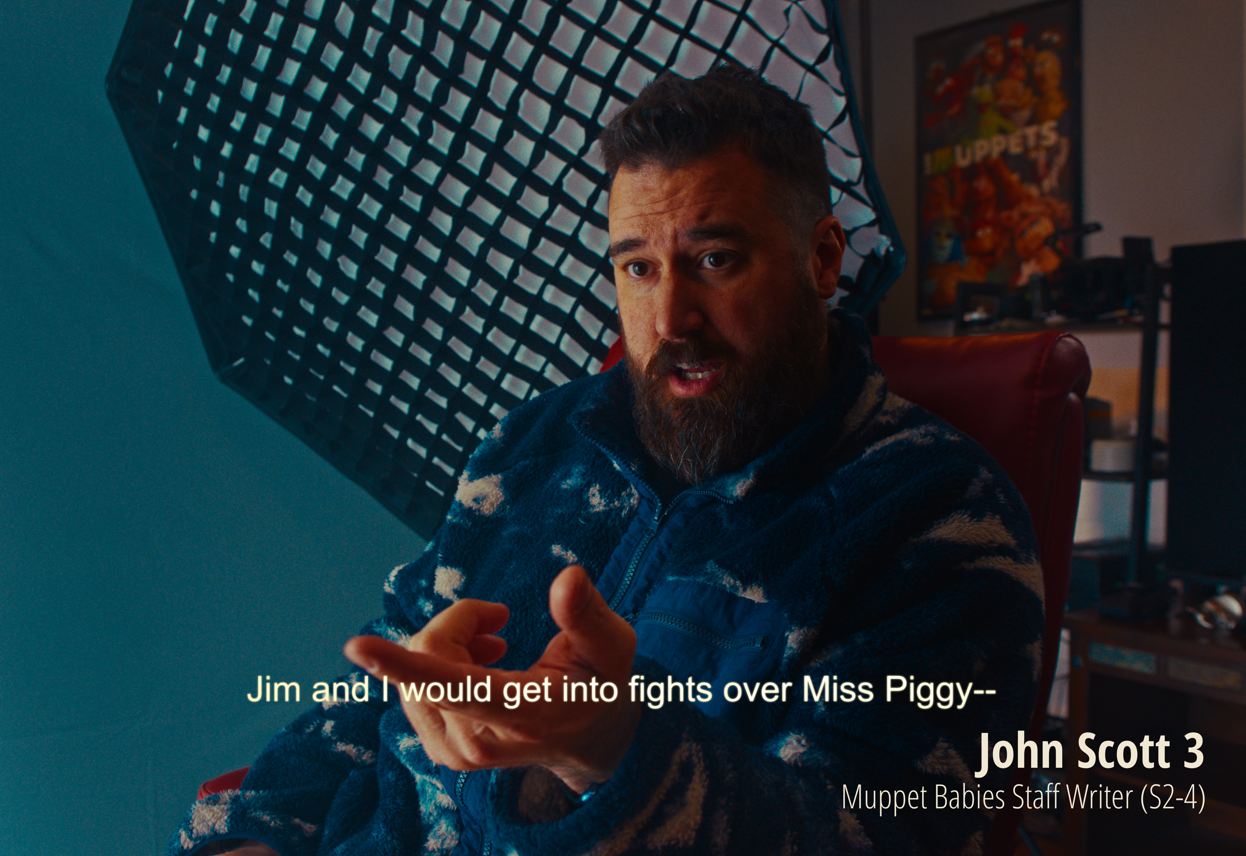 A man with a beard and short hair, wearing a blue and beige patterned fleece, is sitting in a room with a red chair and computer equipment. Behind him, there is a large light diffuser and a poster of the Muppets. The man is gesturing with his hands a