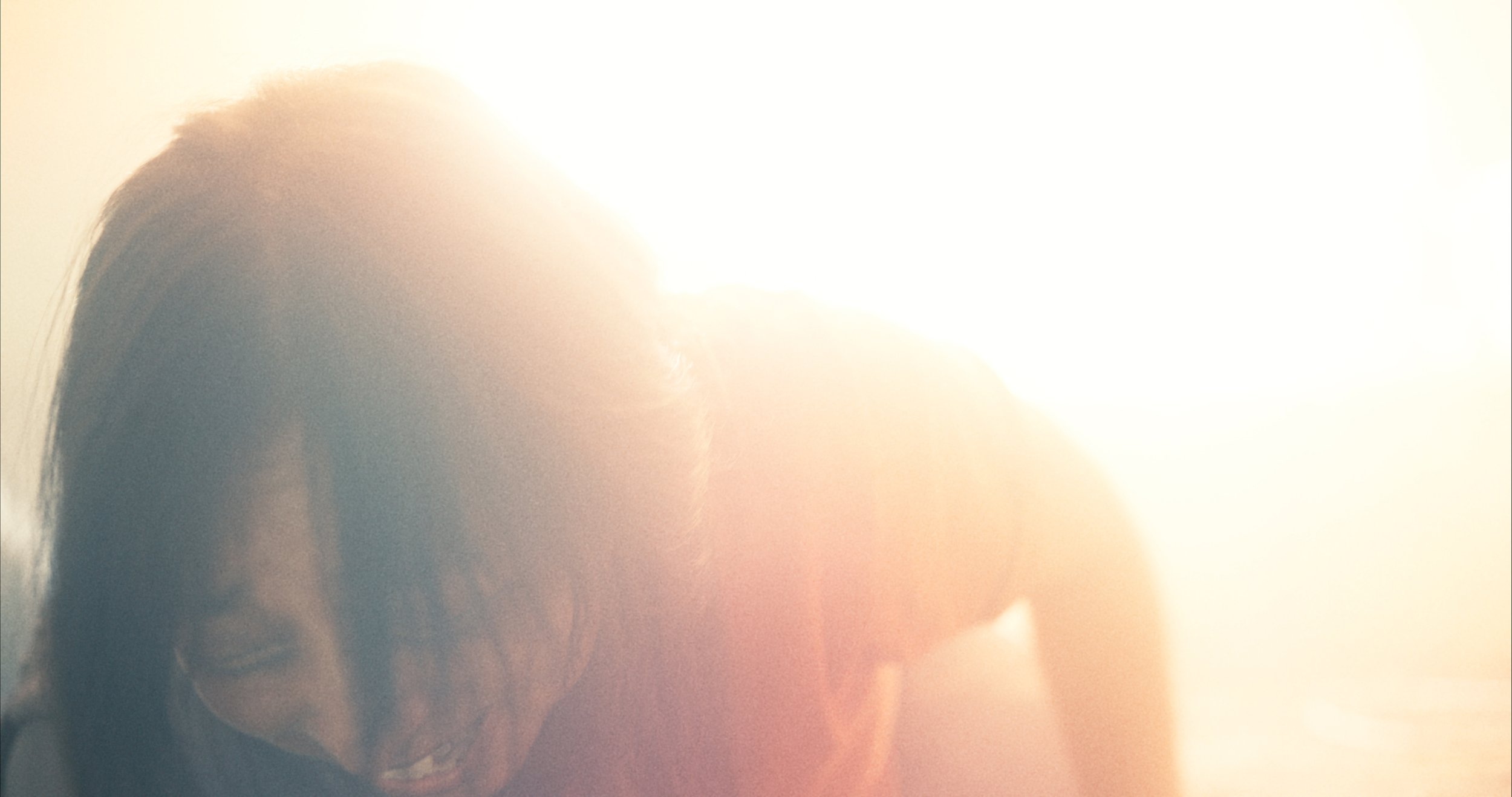 A woman with dark hair smiling in front of bright sunlight, with the light causing overexposure in the photo.