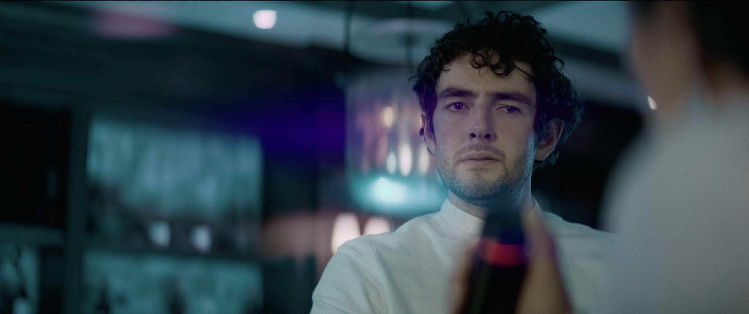 A man with curly hair and a serious expression looking at a soda bottle in the foreground in a dimly lit space.