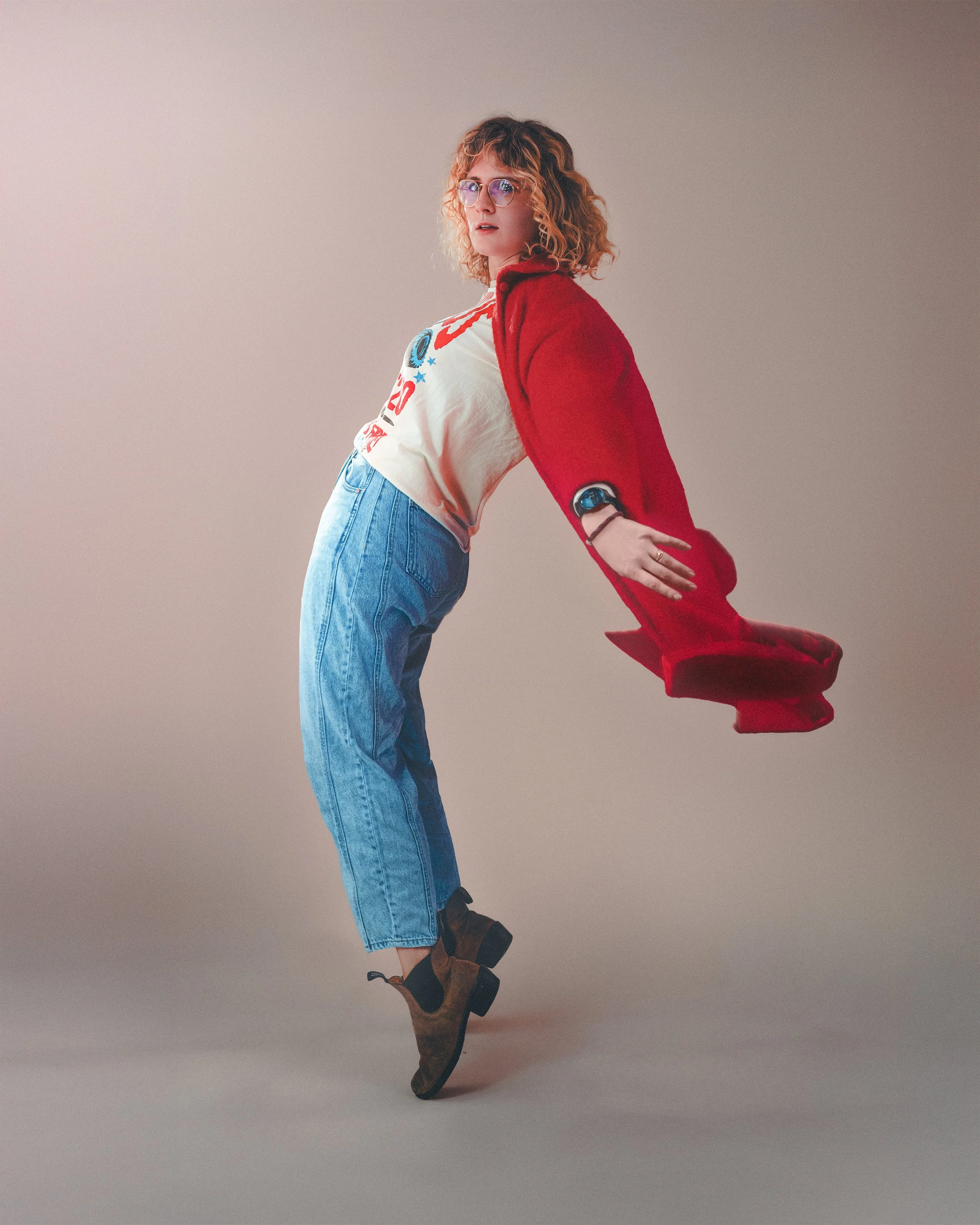 A woman with curly red hair and glasses is standing with her upper body leaning back, wearing a red jacket, a graphic t-shirt, blue jeans, and brown boots, against a plain background.