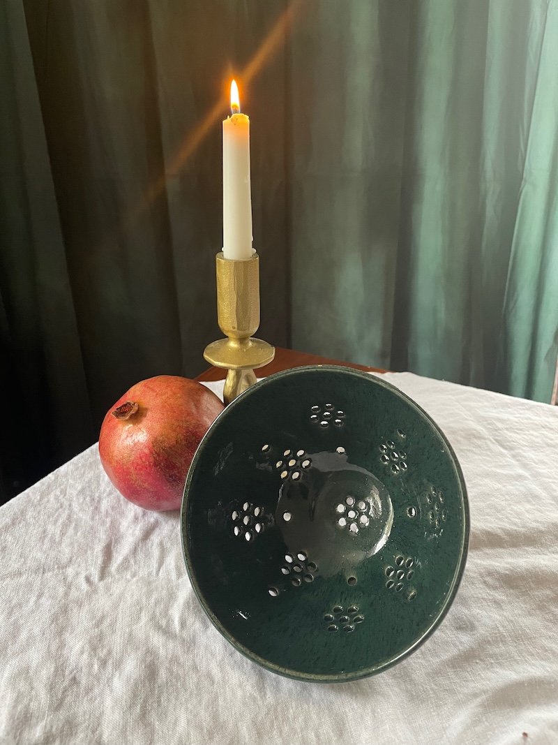 Dark green bowl with colander holes before a lit candles and a pomegranate RVA