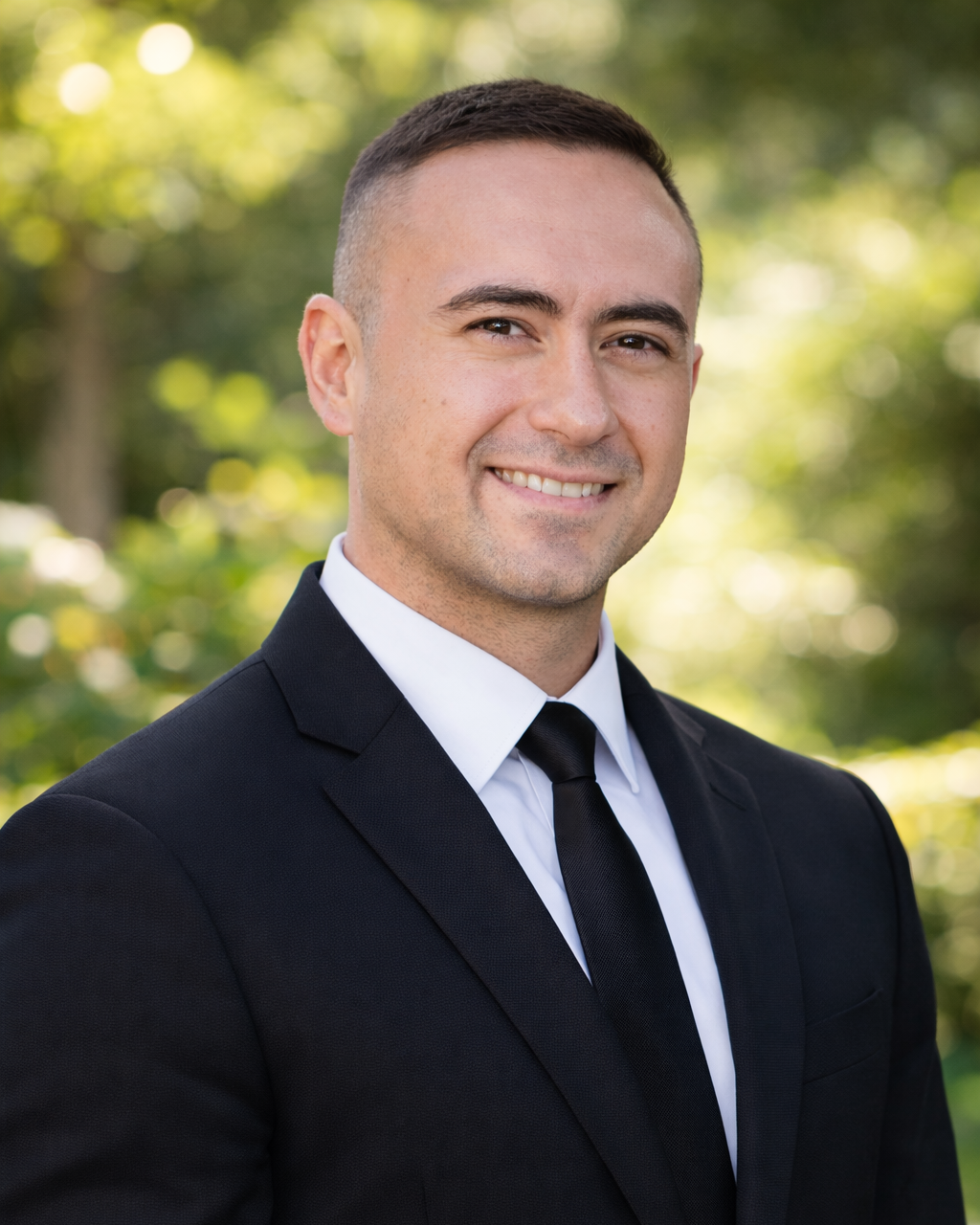 Portrait of a smiling man wearing a black suit, white shirt, and black tie, outdoors with blurred green foliage in the background.
