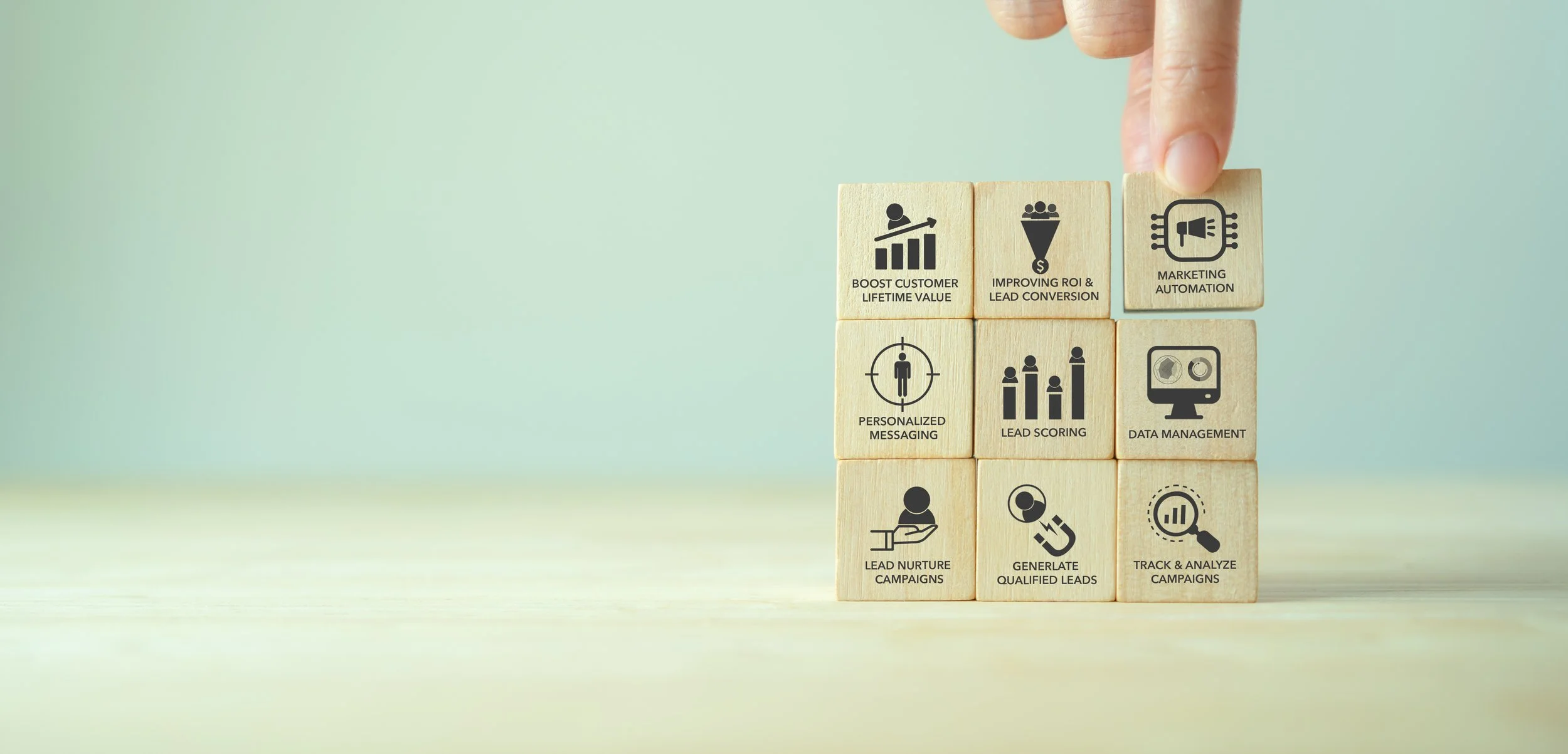 A hand pointing to wooden blocks with marketing icons and labels, including boosting customer lifetime value, improving ROI & lead conversion, marketing automation, personalized messaging, lead scoring, data management, lead nurture campaigns, generating qualified leads, and tracking & analyzing campaigns.