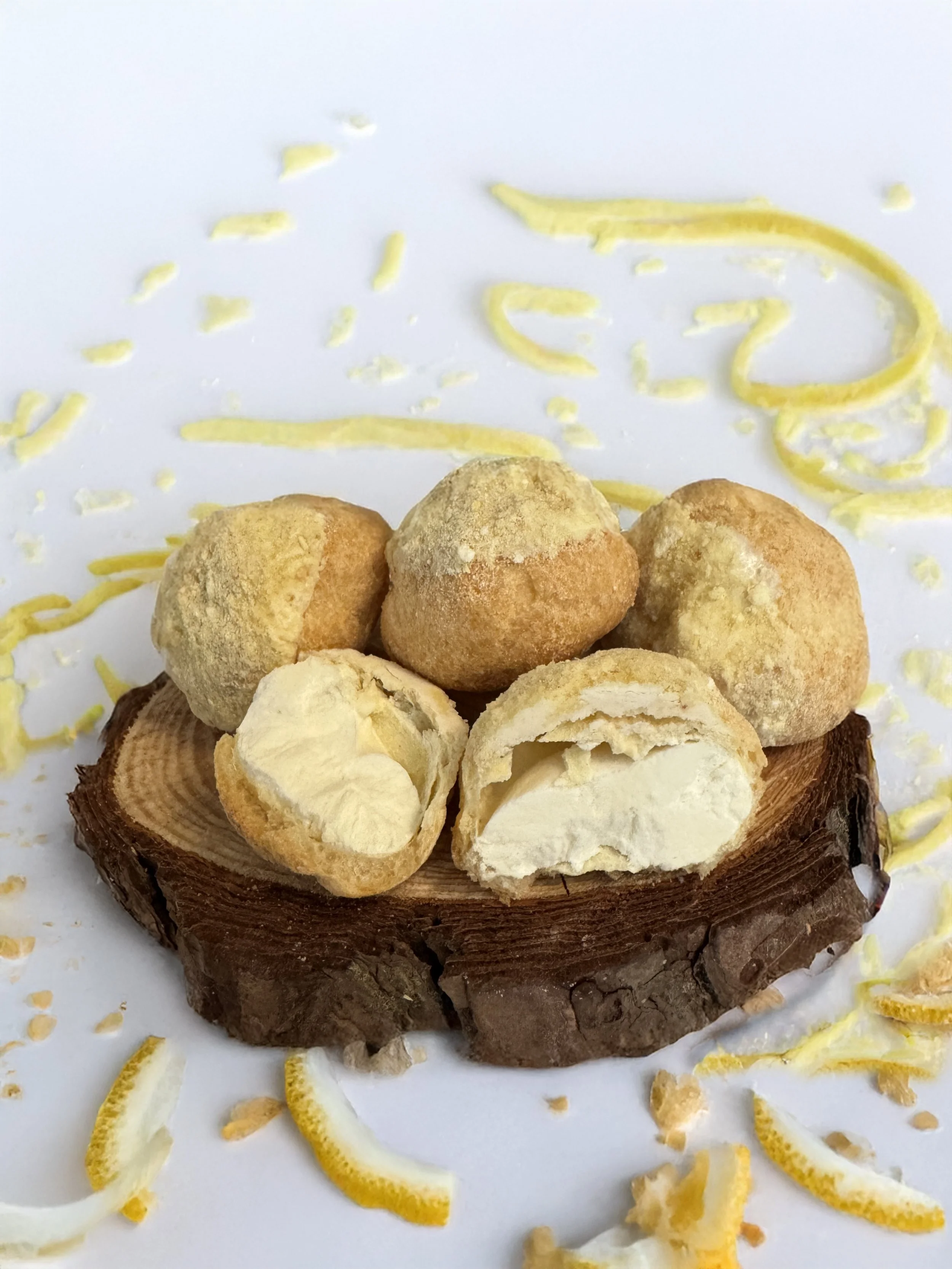 Freeze dried Lemon Cream Puffs — airy vanilla-filled pastry bites coated in lemon powder, displayed on a wood slice with lemon zest pieces scattered on a white background