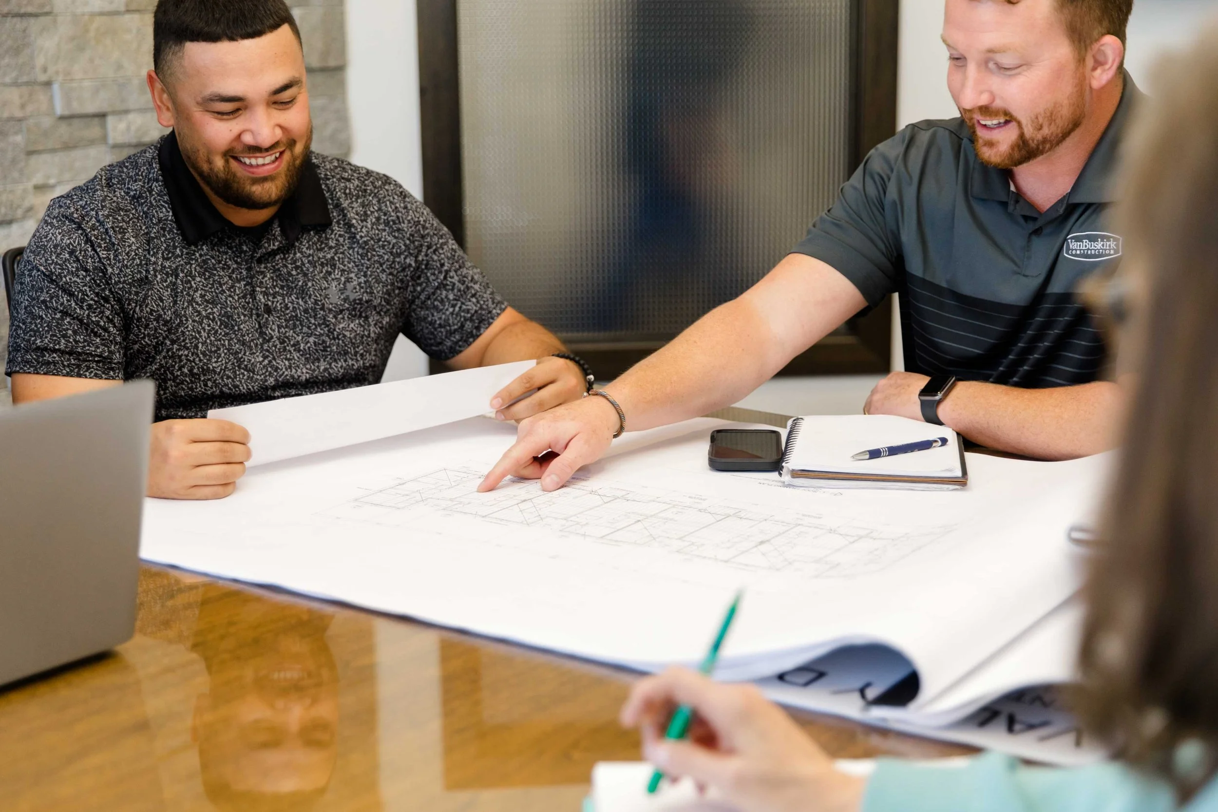 Two men looking at architectural blueprints on a table, one of them is pointing at a specific area.