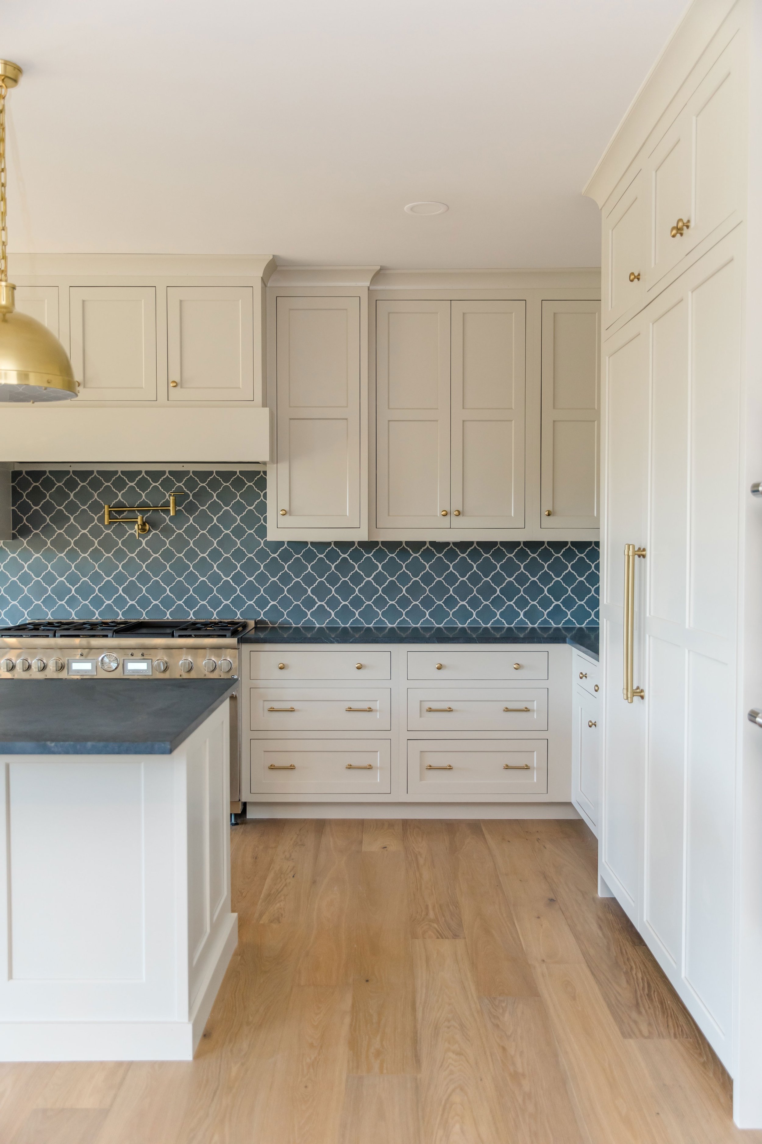 A modern kitchen with white cabinetry, a blue moroccan tile backsplash, and gold hardware, featuring a stainless steel stove and light hardwood floors.