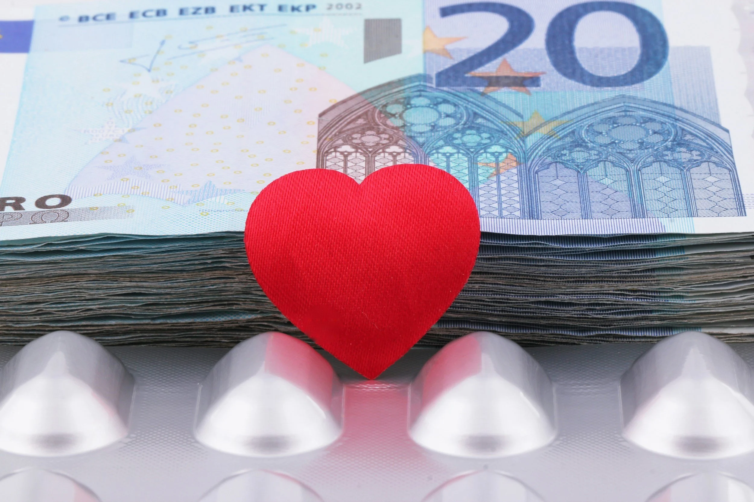 A stack of 20 euro banknotes, a red heart, and white pills in a blister pack.